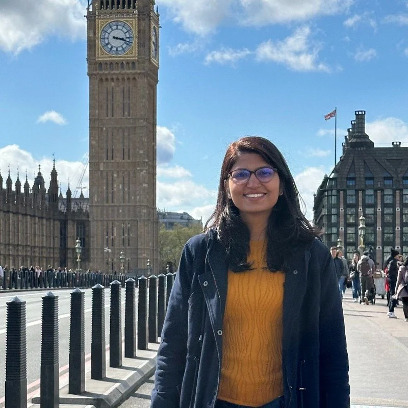 A woman with glasses and dark hair smiling in front of Big Ben in London, wearing a yellow sweater and dark jacket, with a cloudy sky and city buildings in the background.