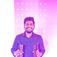 A man with a beard smiling holding two trophies against a pink background with white dots.