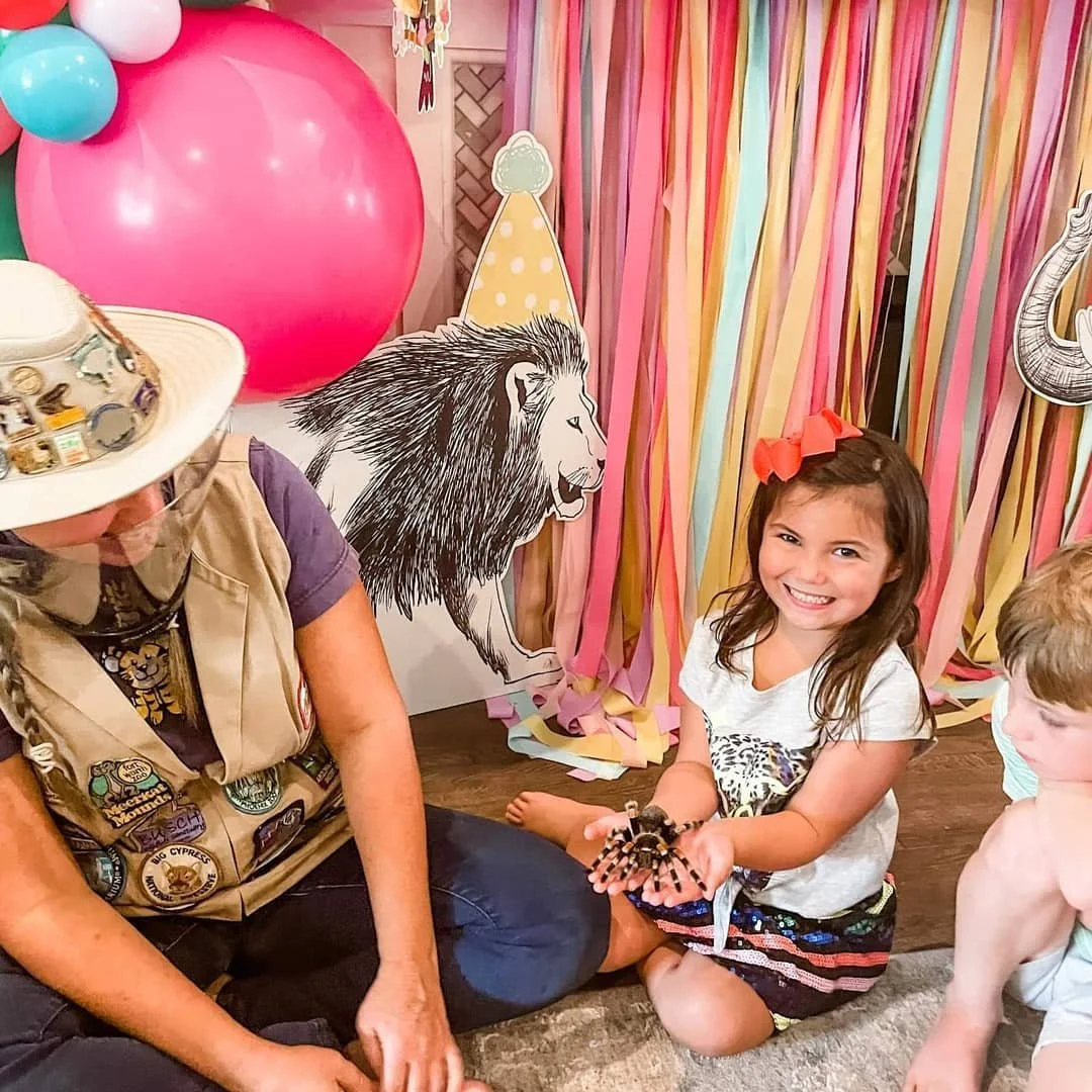 Young girl smiling and holding a spider at a birthday party with colorful streamers and lion decoration in the background.