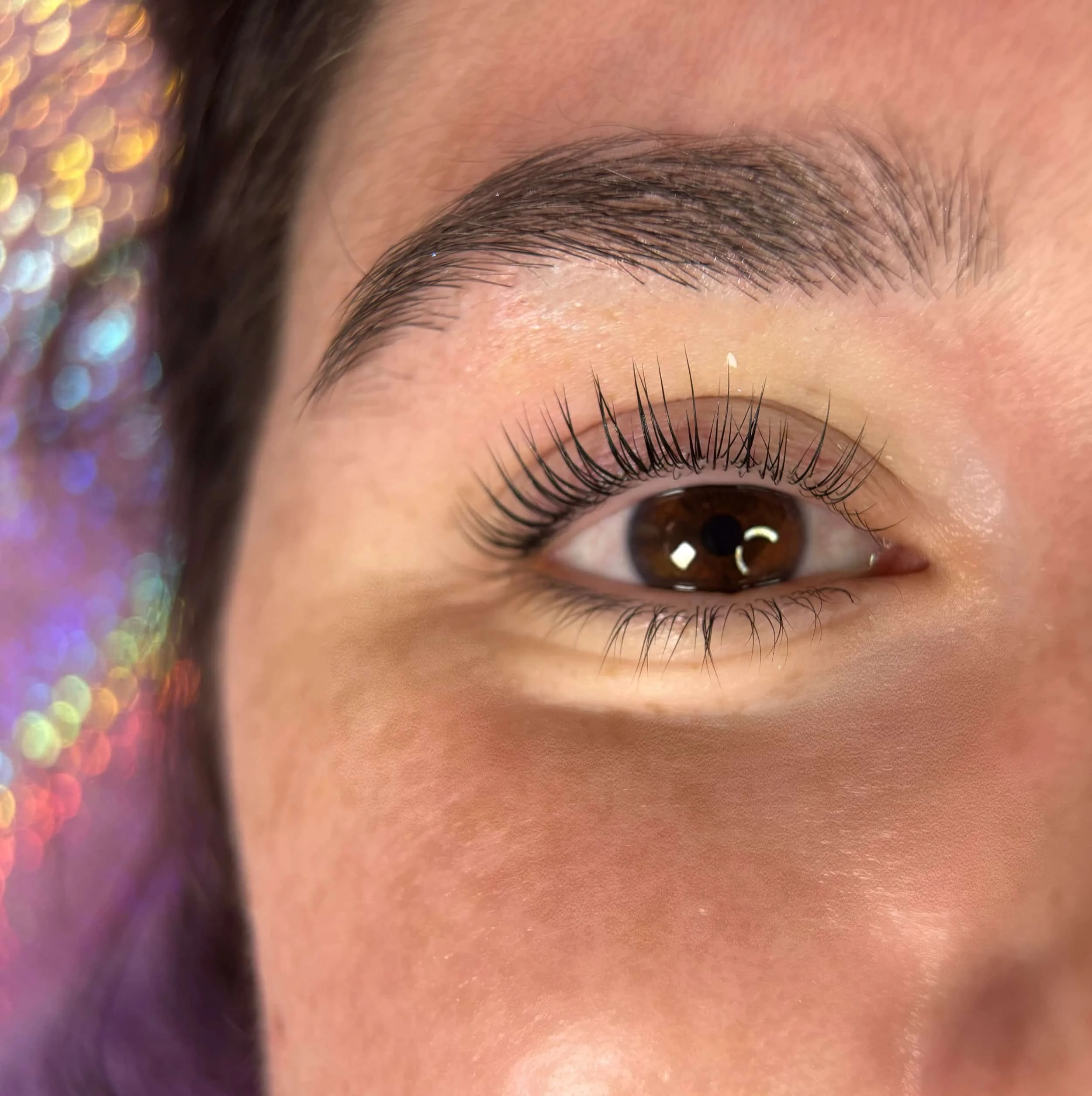 Lähellä kuvan tarkka silmän ja kulmakarvan lähikuvasta, jossa näkyy ruskea silmä, pitkät ripset ja siisti kulmakarva, taustalla värikkäitä valoja.