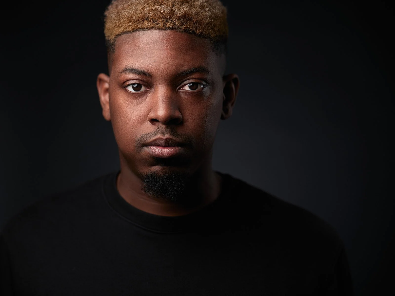 Close-up portrait of a young man with black curly hair dyed blonde on top, wearing a black shirt, against a dark background.