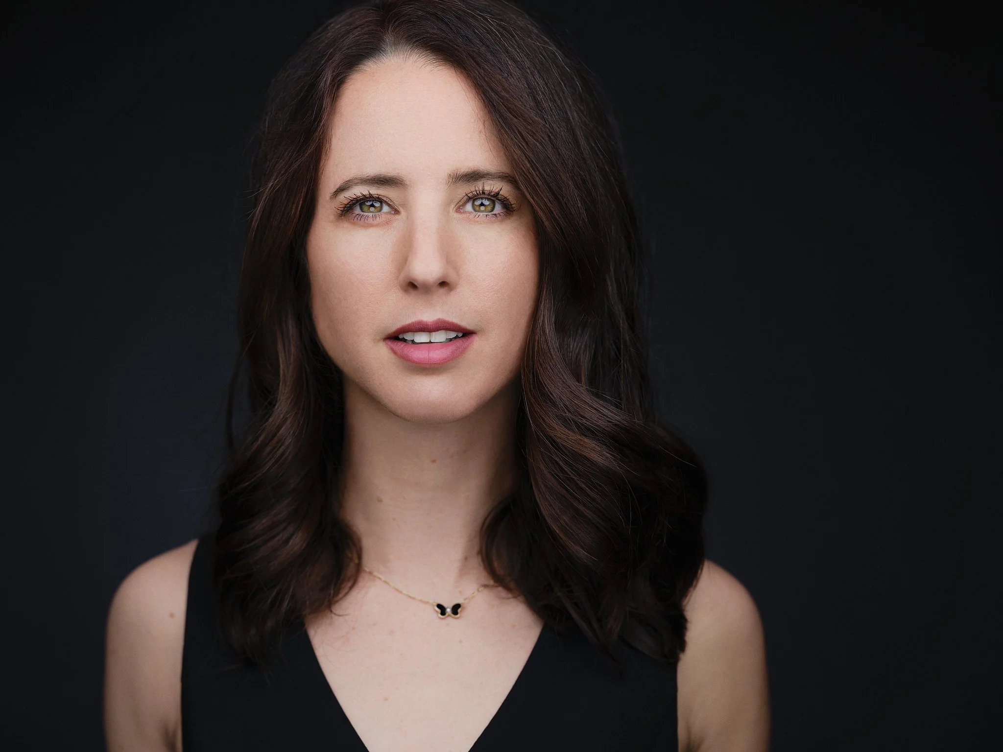 Portrait of a woman with shoulder-length dark brown hair, light skin, light-colored eyes, wearing a black sleeveless top and a delicate necklace, against a dark background.