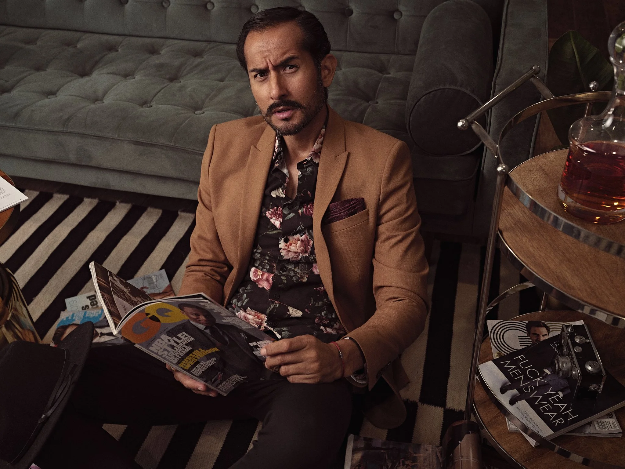 A man with dark hair and a beard, wearing a tan blazer and floral shirt, is sitting on the floor with a magazine in his hand, looking confused or surprised. He is in a living room with a gray sofa, patterned carpet, and a wooden coffee table with mag