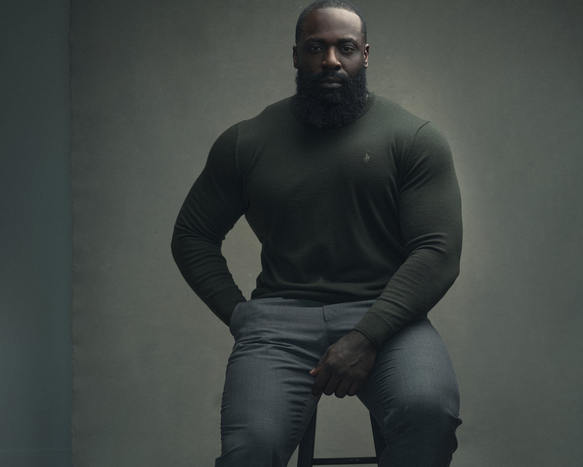 A man sitting on a stool against a gray background, wearing a dark green long-sleeve shirt and gray pants.