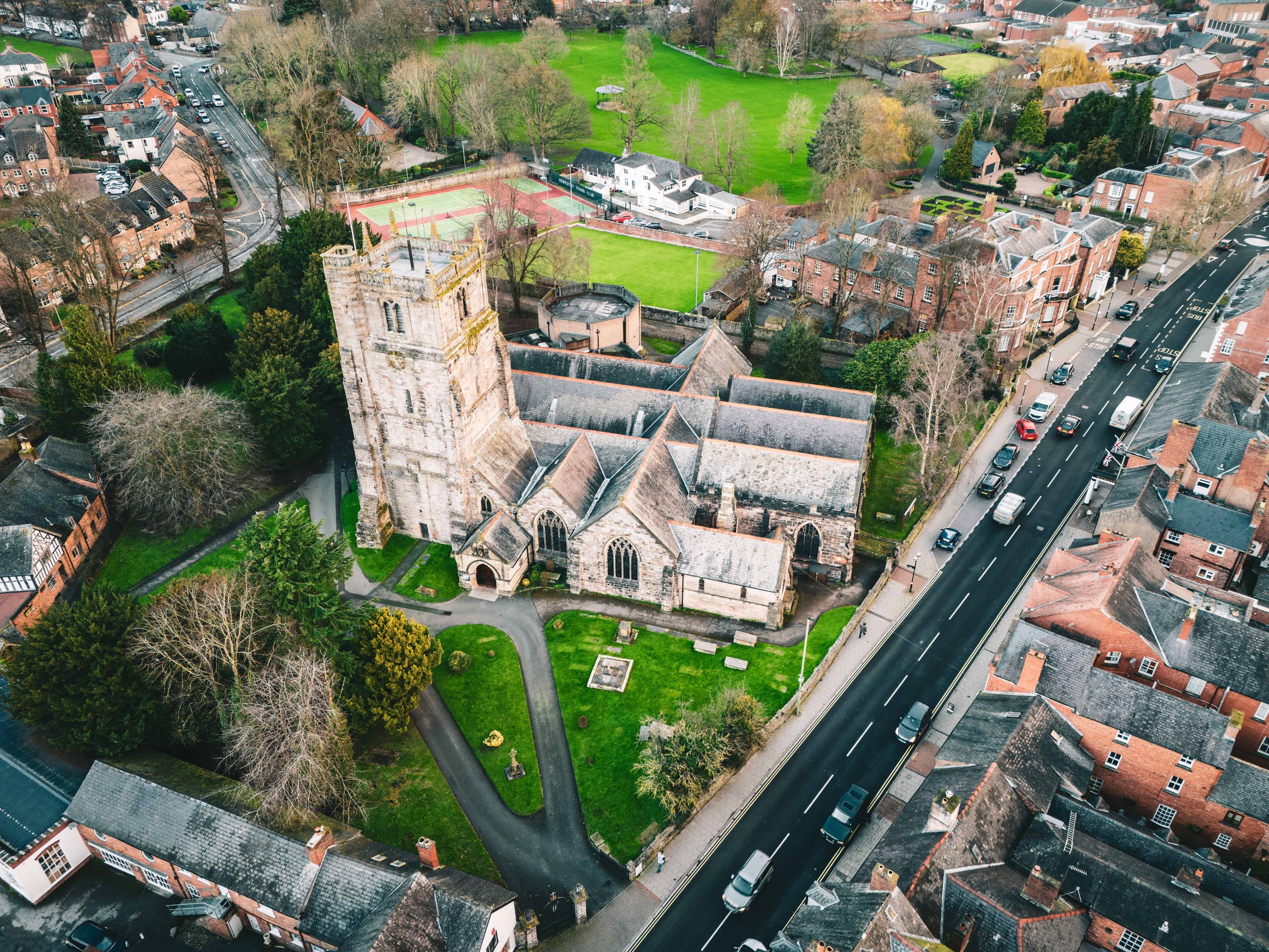 Aerial view of a historic church with a tall tower in an English town, surrounded by greenery, roads, and residential buildings.