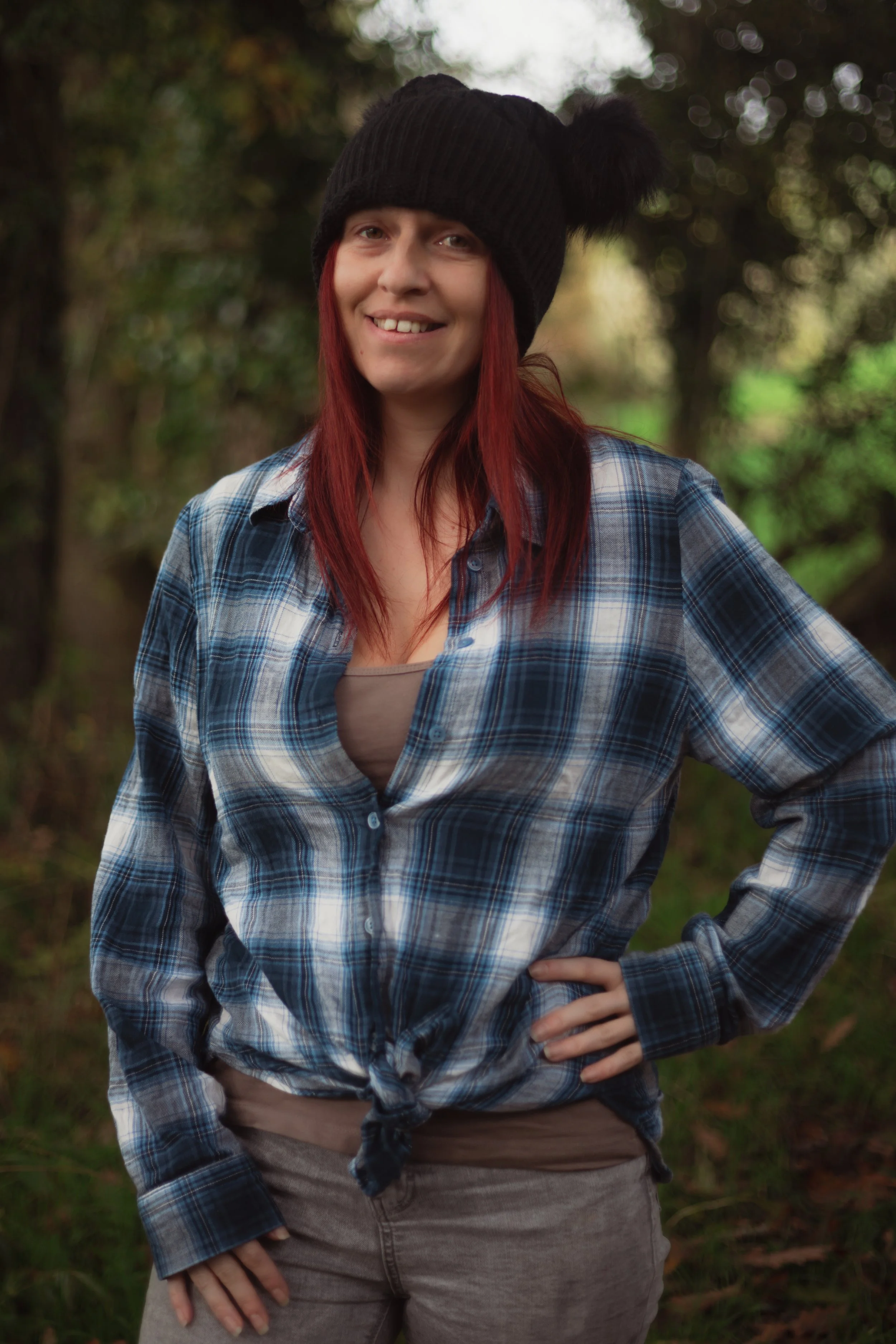 A woman with red hair wearing a black knit hat with pom-poms, a plaid shirt tied at the waist, and gray pants, standing outdoors in a wooded area.