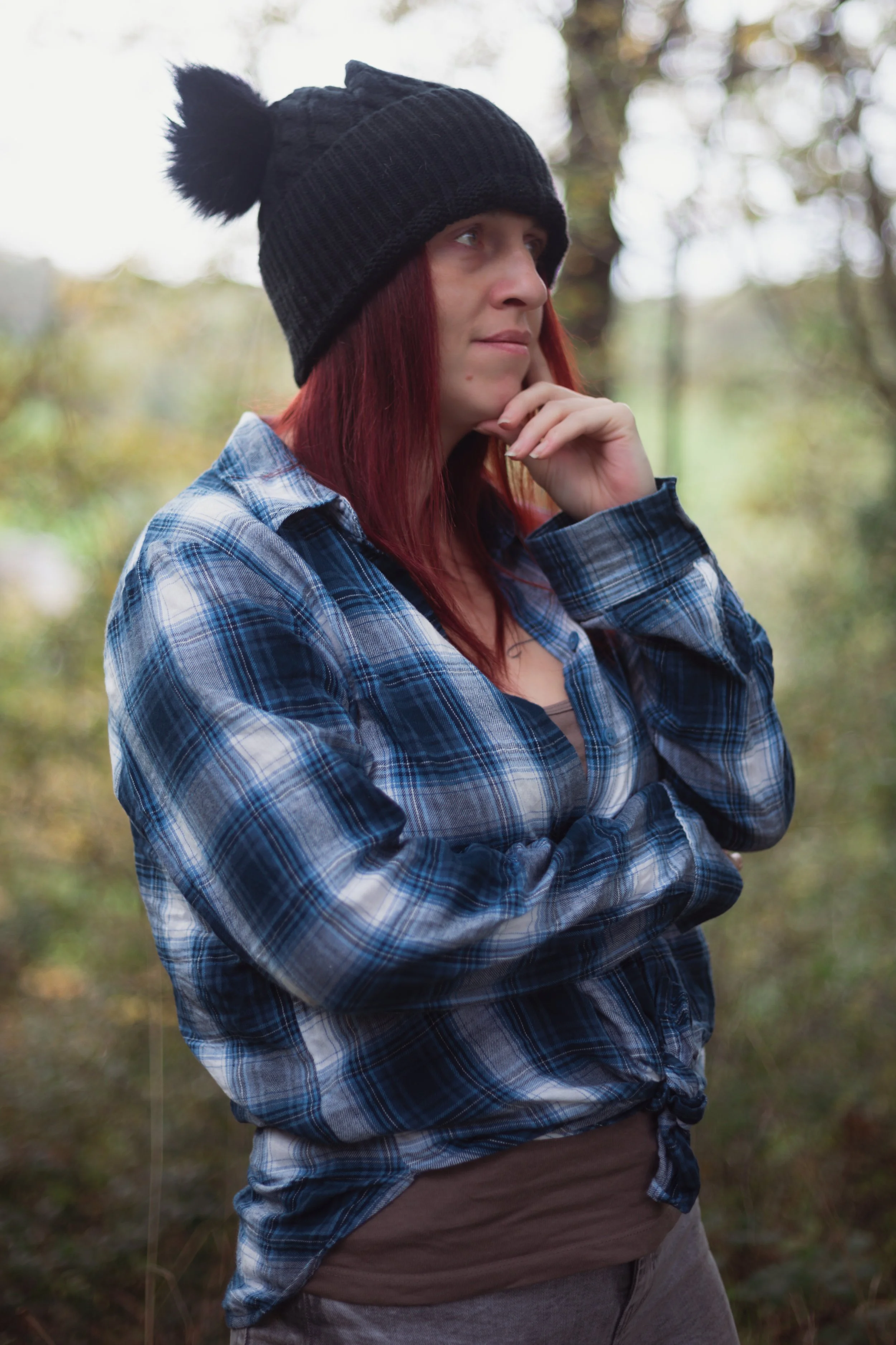 A woman with red hair wearing a black knit hat with a pom-pom, a blue plaid shirt, and gray pants, standing outdoors among trees, with a thoughtful expression.