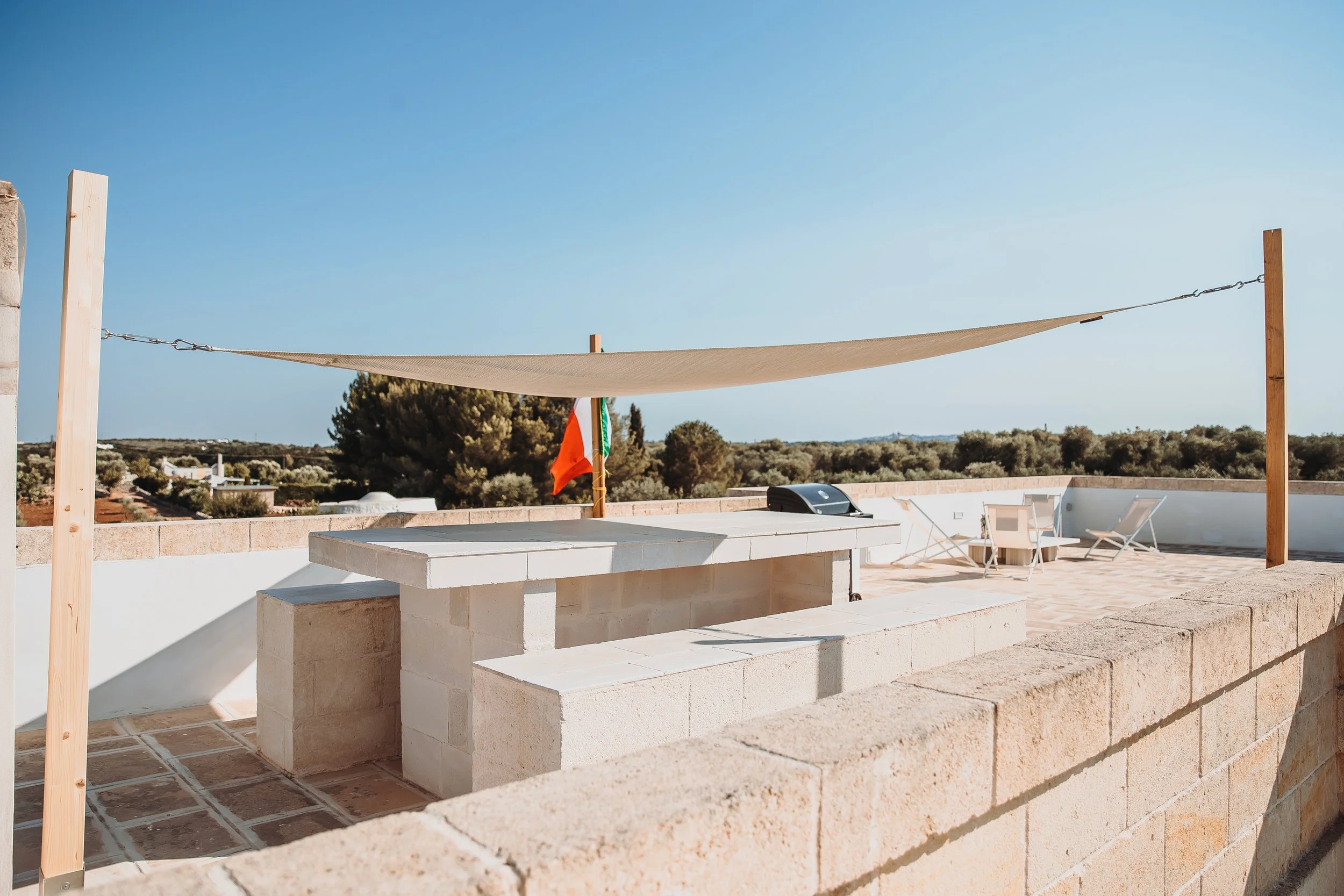 Rooftop patio with outdoor seating, a barbecue, and a shade sail, overlooking a landscape with trees and a clear blue sky.