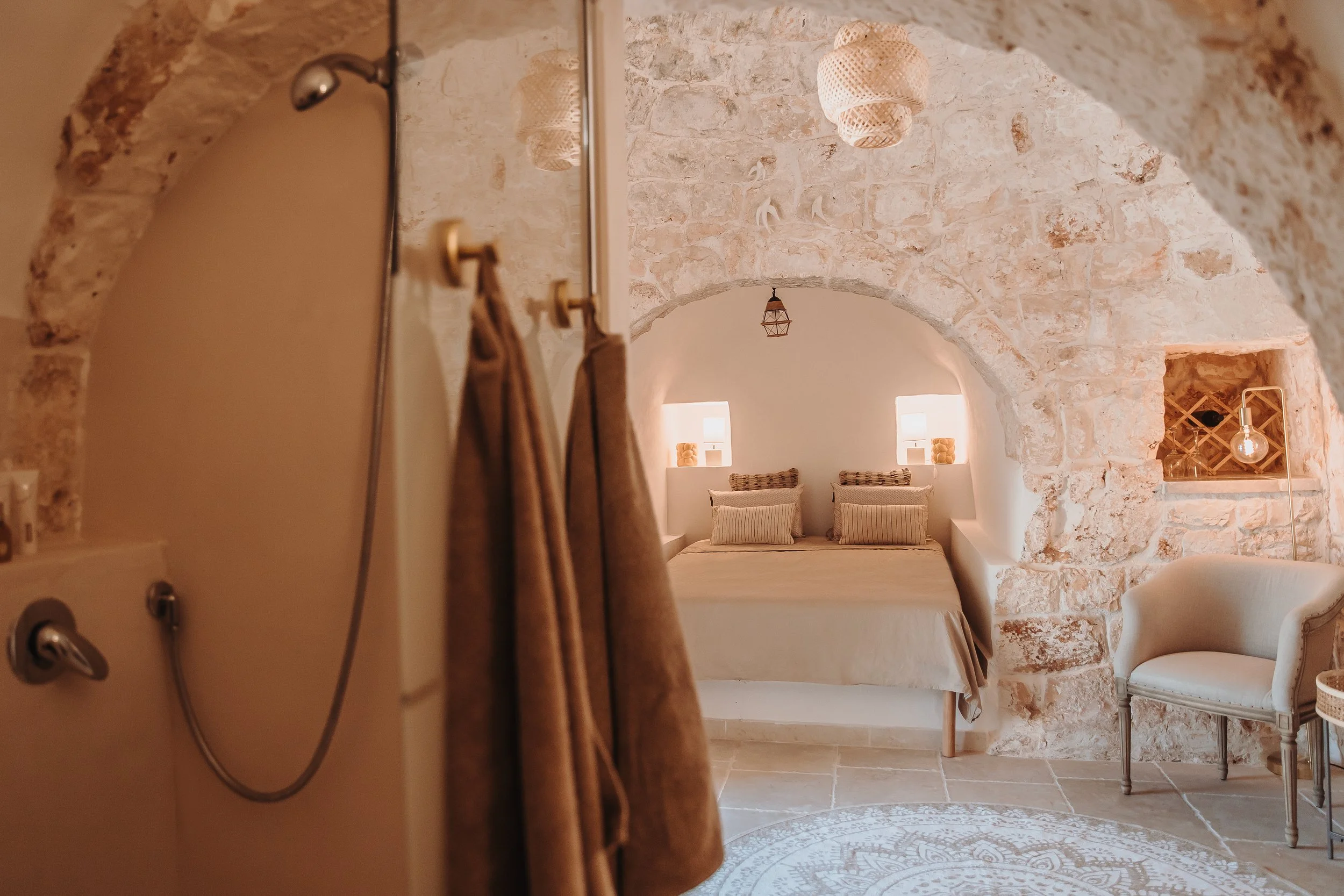 Interior of a cozy bedroom with stone walls, arched ceiling, bed with striped pillows, small windows with lamps, and a seating area with a white upholstered chair. A partial view of a shower with a showerhead and brown towels hanging.