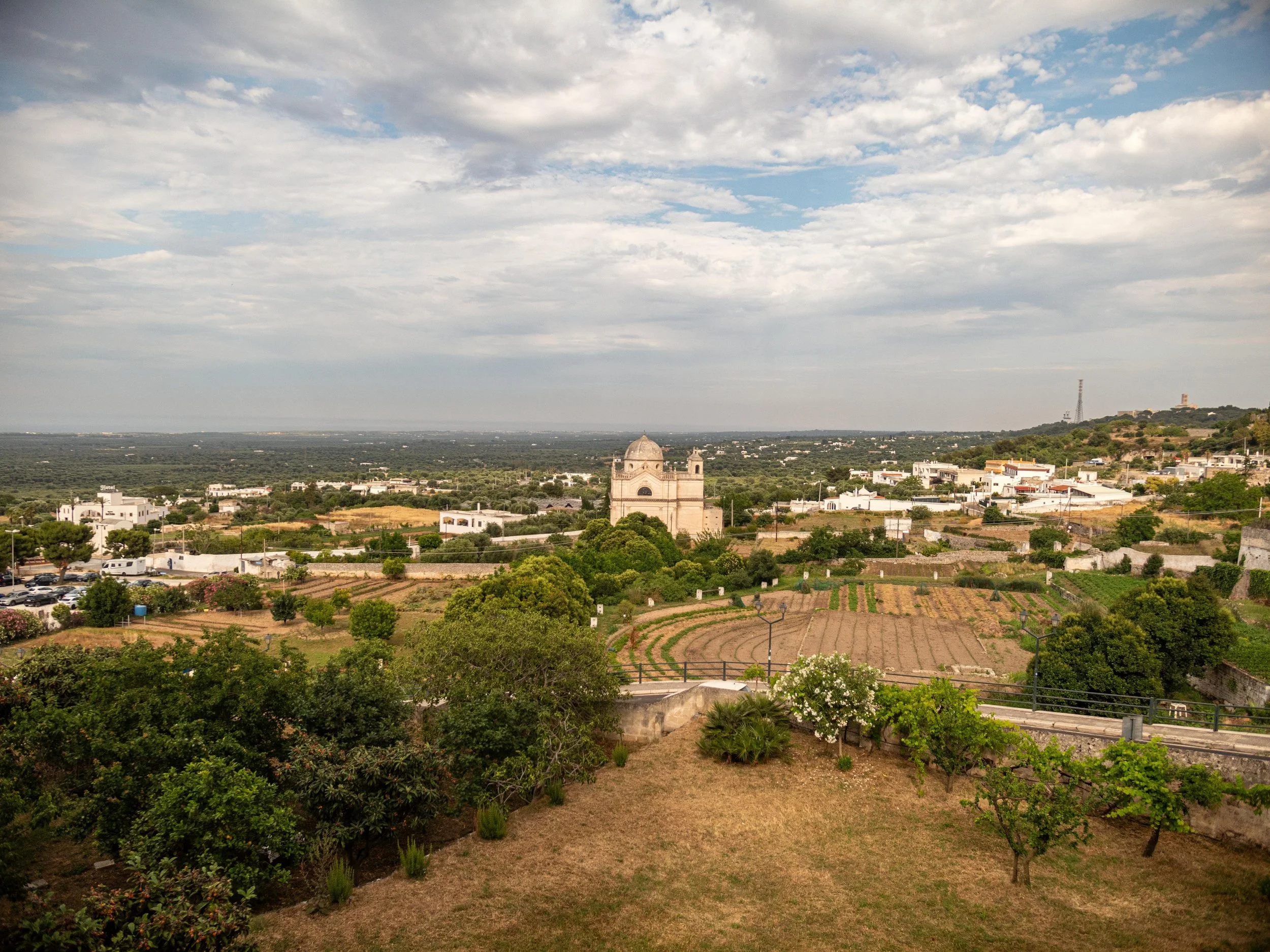 Landscape view of the countryside of Ostuni with a historic church in the center, surrounded by trees, farmland, and rolling hills, under a partly cloudy sky.