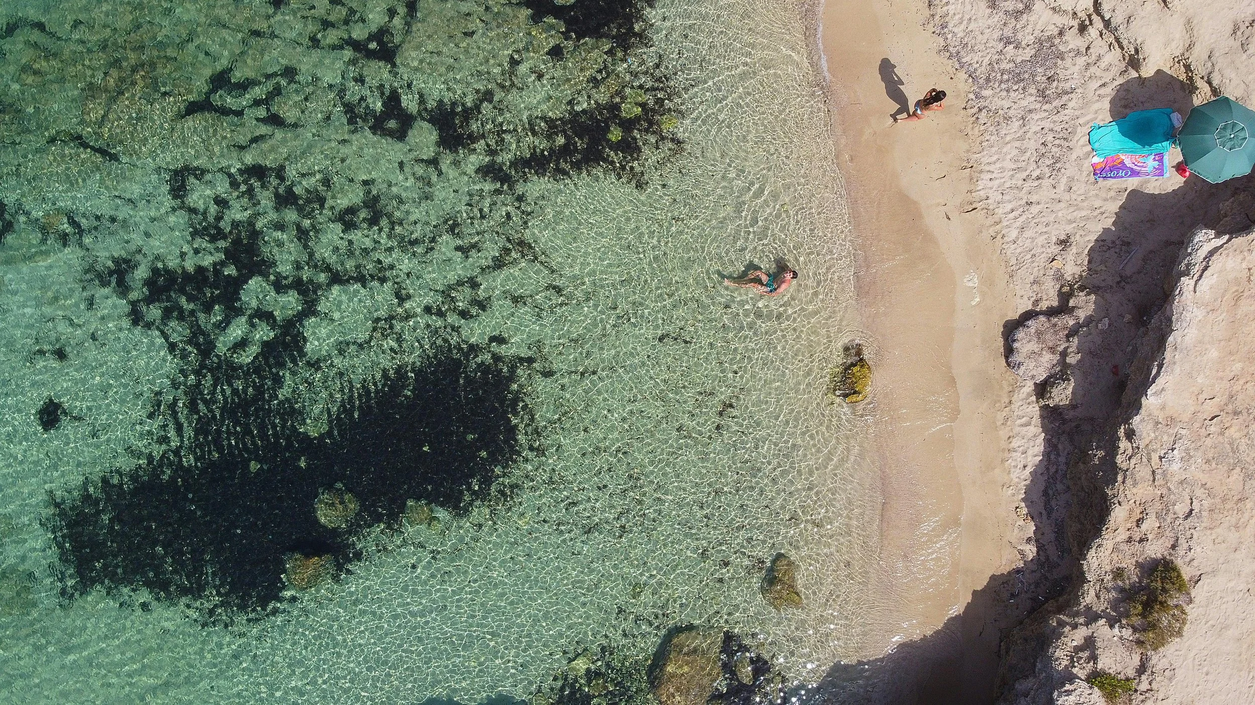 Aerial view of a beach with clear water, rocks, and a sandy shore. There are three people in the water, and a small group of people on the beach near an umbrella and colorful towel.