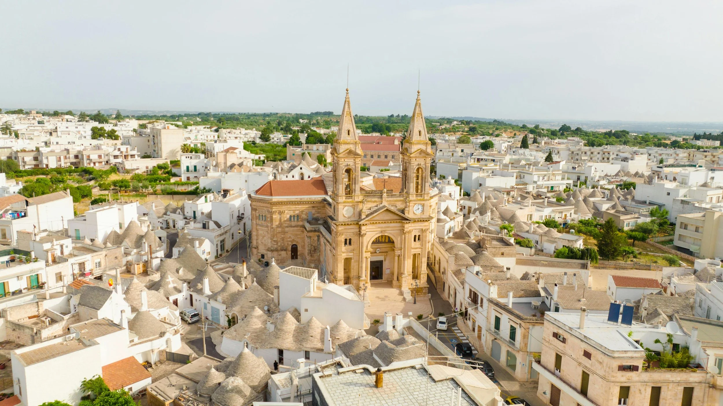 Aerial view of a historic church with twin steeples in a white-washed town with many small buildings and rooftops, some conical, with green trees scattered throughout.