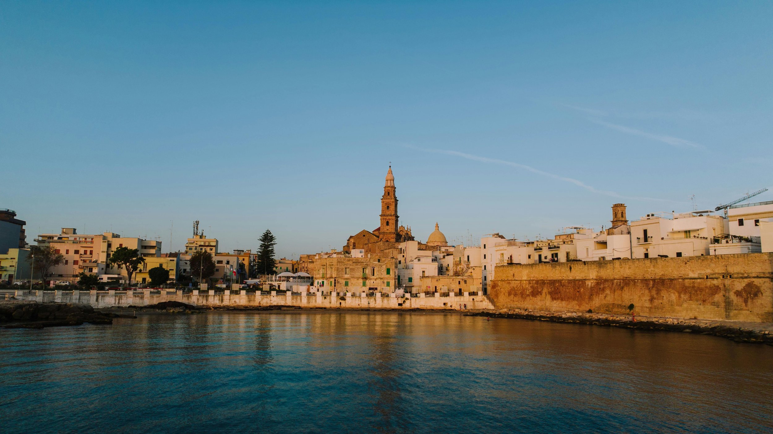 View of a historic cityscape with a church tower and domed building along a waterfront at sunset.