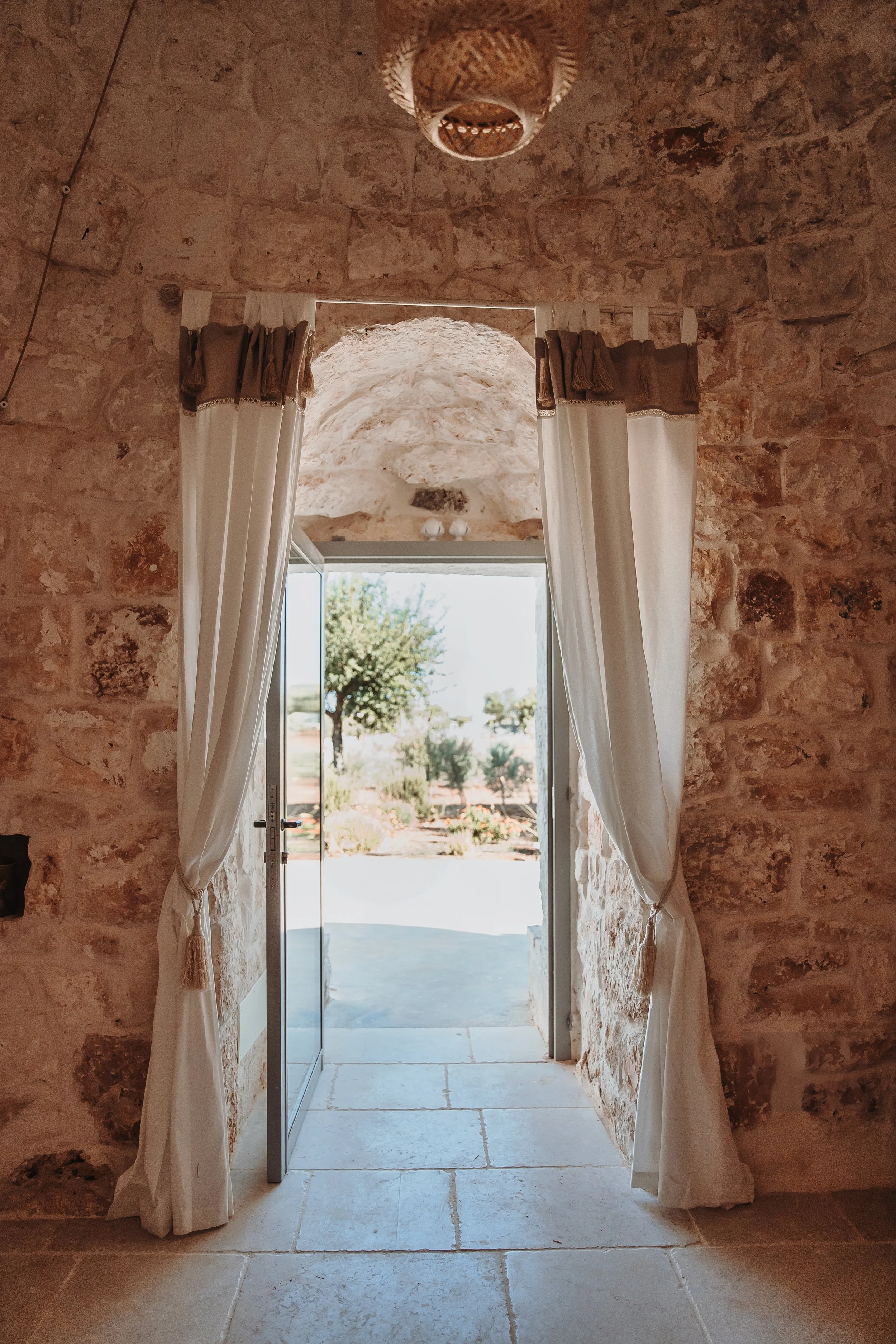 Open stone doorway with white curtains opening to an outdoor landscape with trees and shrubs.