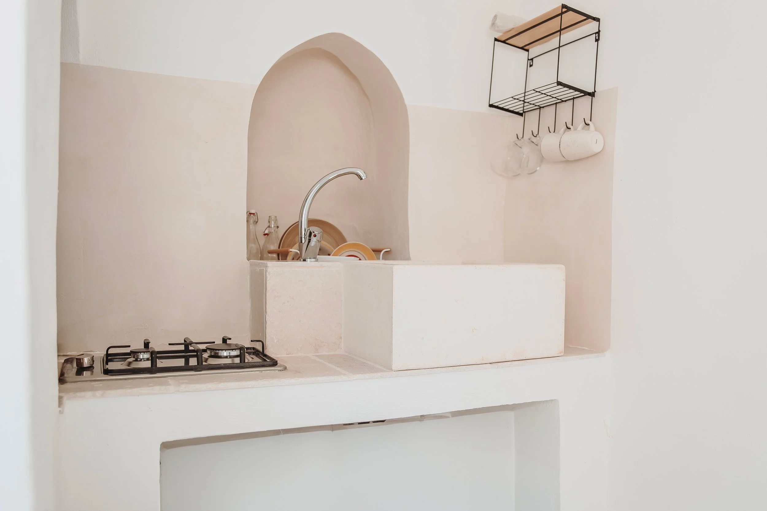 Minimalist kitchen with a gas stove, a small sink with a curved faucet, and a wall-mounted metal rack holding glasses, mugs, and a dishcloth.