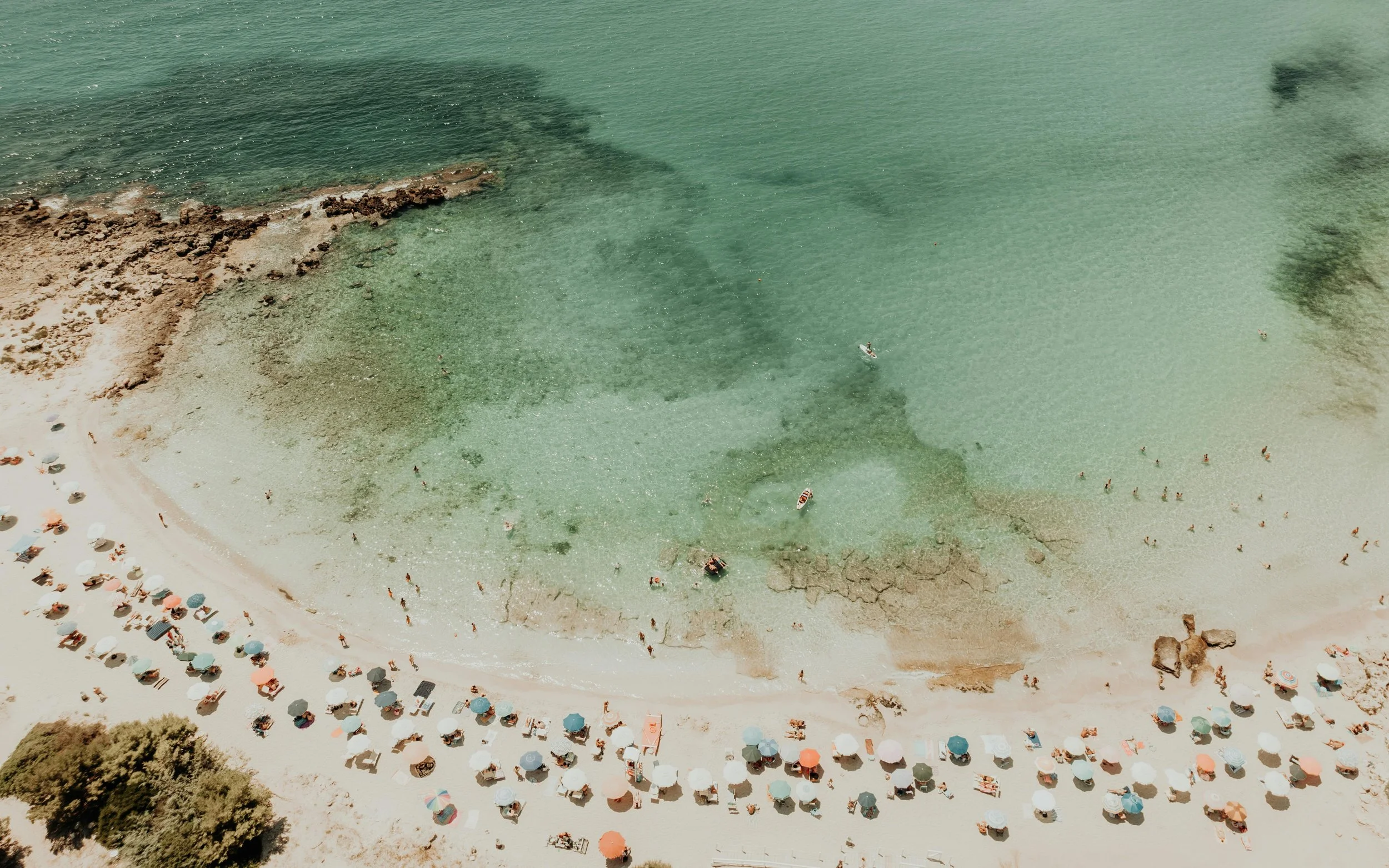 Aerial view of a beach with umbrellas, people, and boats in clear water.