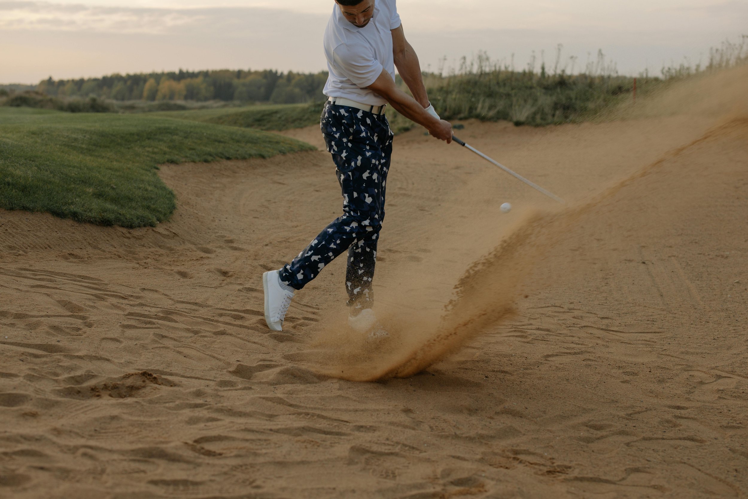 Person hitting a golf ball out of a sand trap on a golf course during sunset.