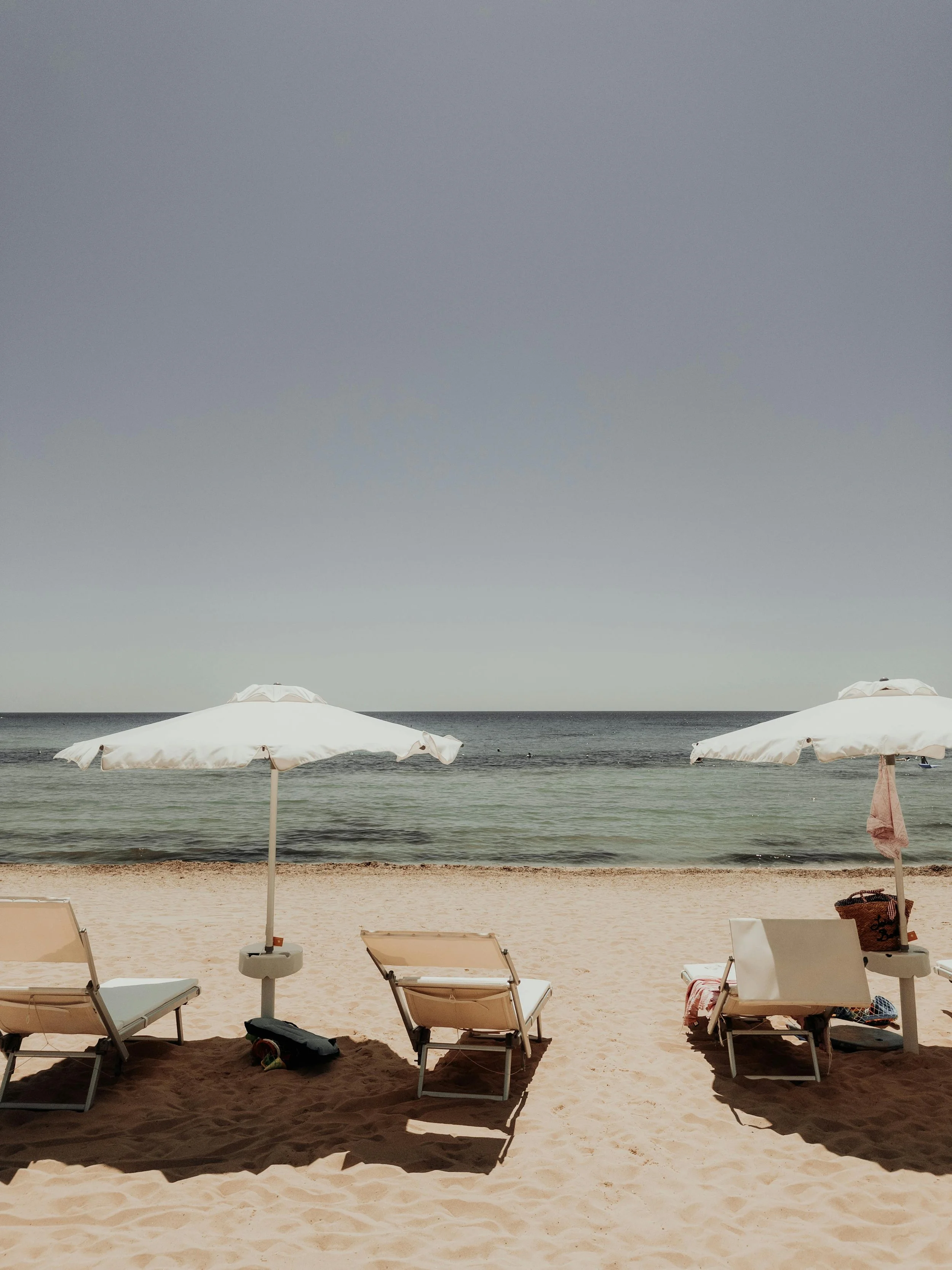 Empty beach scene with two white umbrellas and four lounge chairs on sand, ocean in the background.