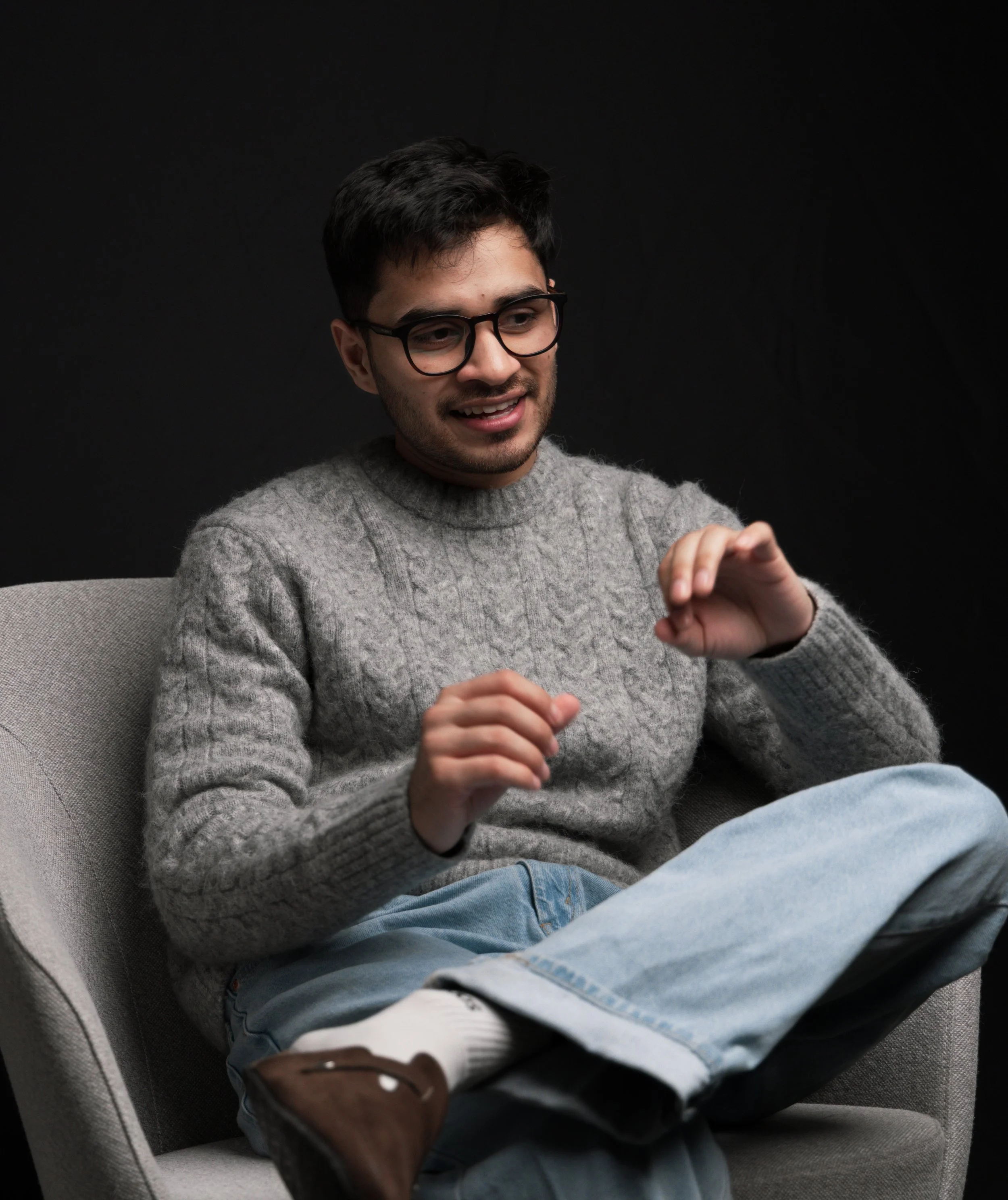 A young man with glasses wearing a gray sweater and light blue jeans sitting on a light-colored chair against a black background, smiling and gesturing with his hands.