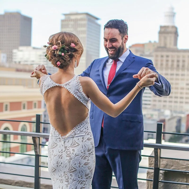 A bride in a lace dress and a groom in a blue suit dancing together on a rooftop with city buildings in the background.
