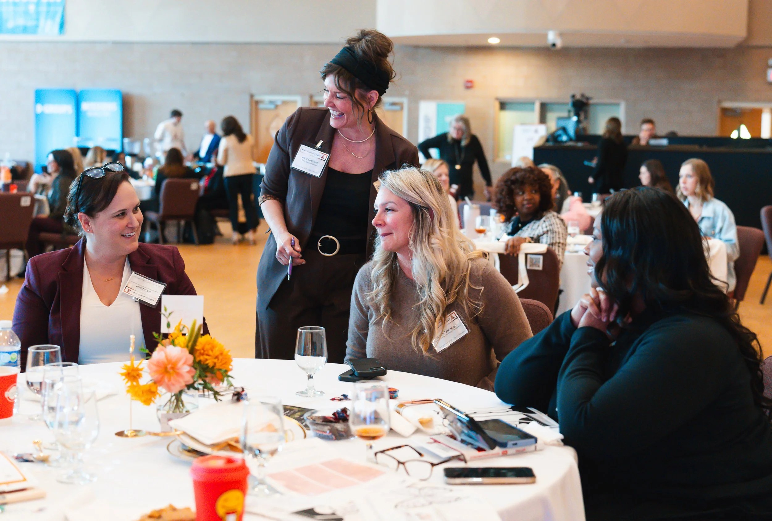 Four women at a conference table having a conversation, with one standing and three sitting, in a large event space with more attendees in the background.