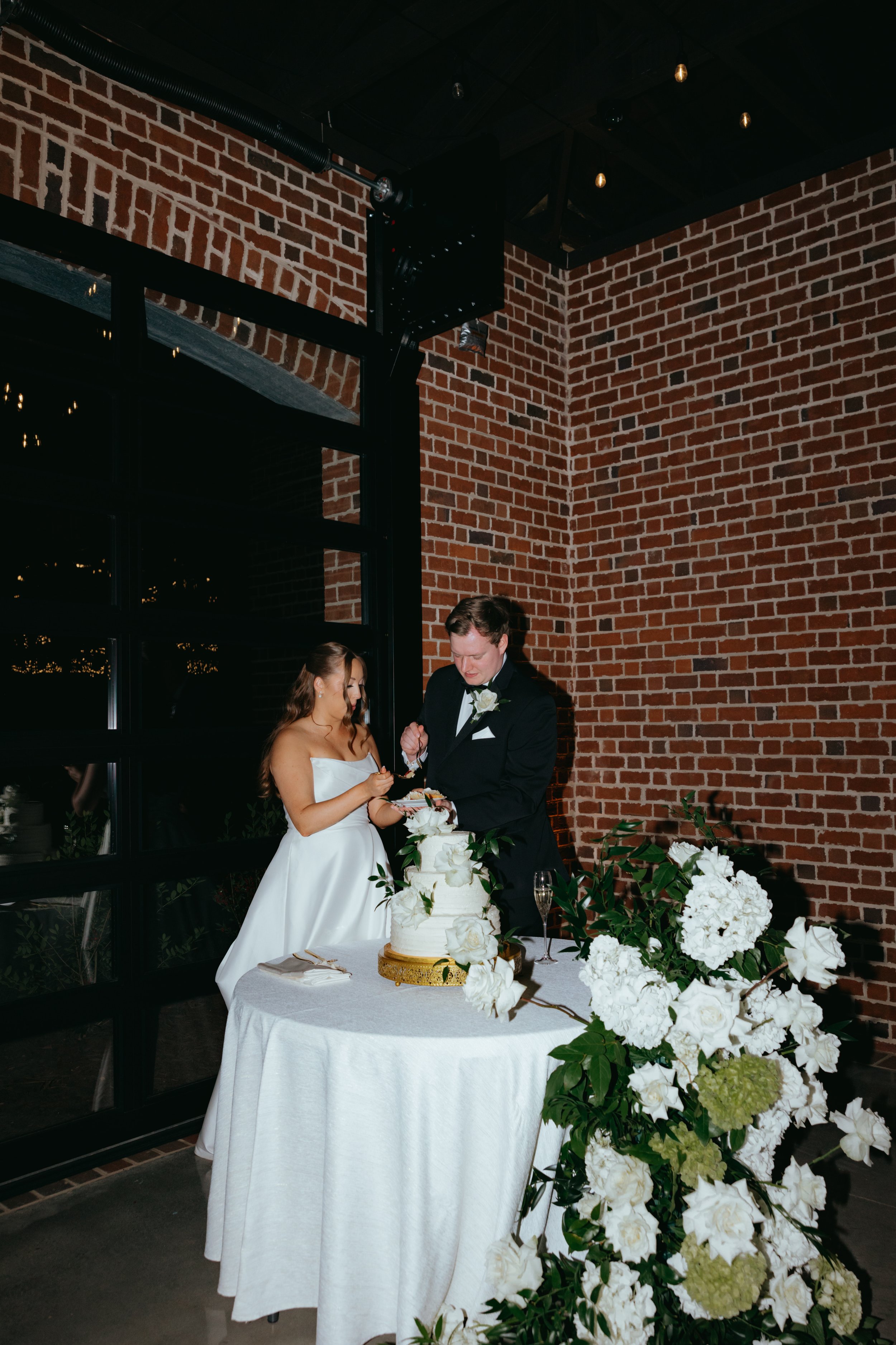 Bride and groom cutting wedding cake at a reception, decorated with white flowers, in front of a brick wall.