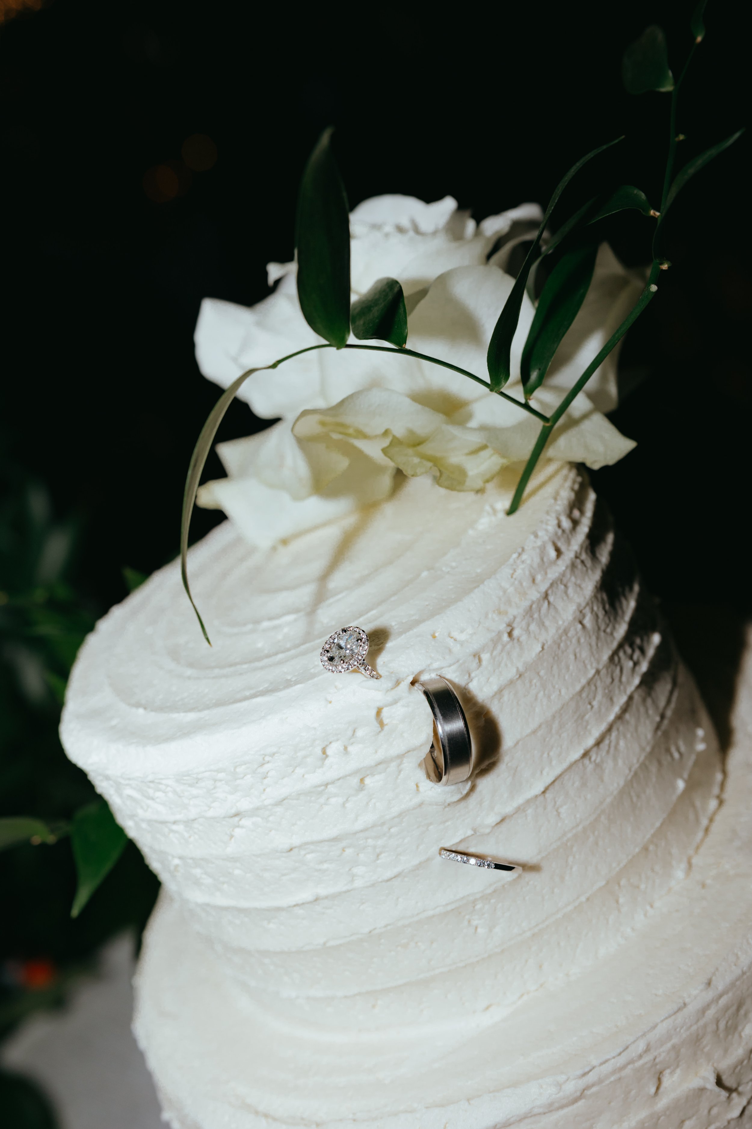 A white frosted cake decorated with white flowers and green leaves, featuring three rings on top.