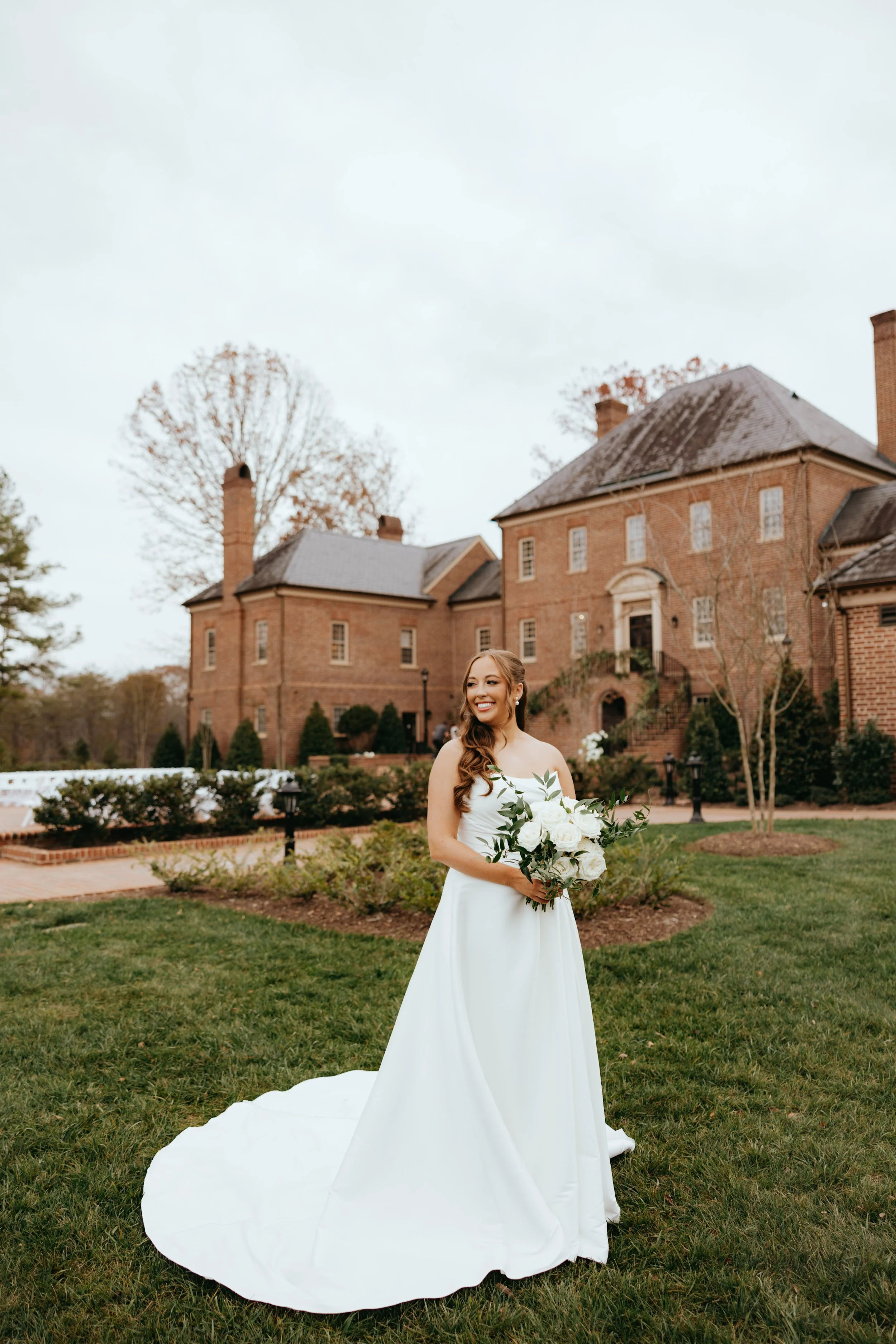 A bride in a white wedding gown holding a bouquet of white roses, standing on a grassy lawn outside a large brick mansion with leafless trees in the background.