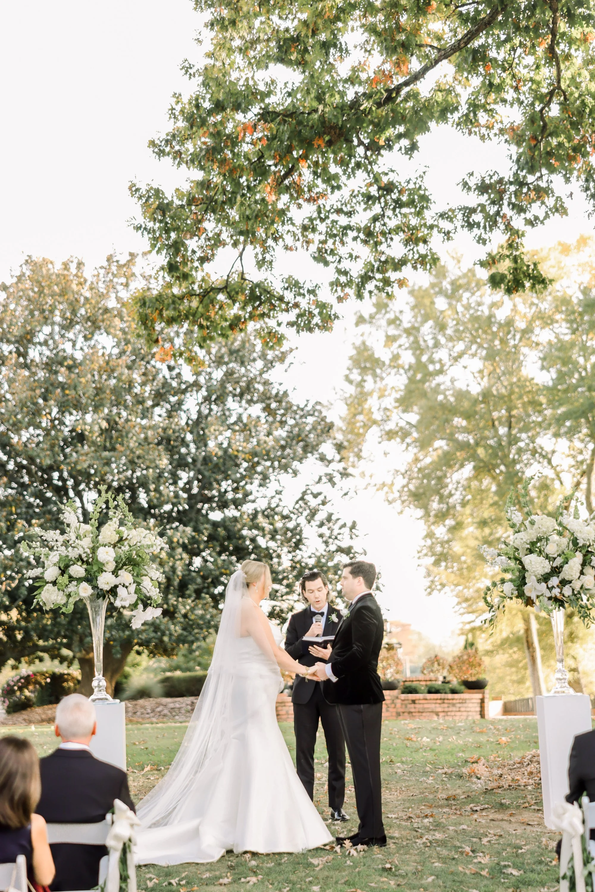 A wedding ceremony outdoors with a bride and groom holding hands and facing each other, an officiant reading from a book, surrounded by guests and large floral arrangements on tall stands, under trees with autumn foliage.