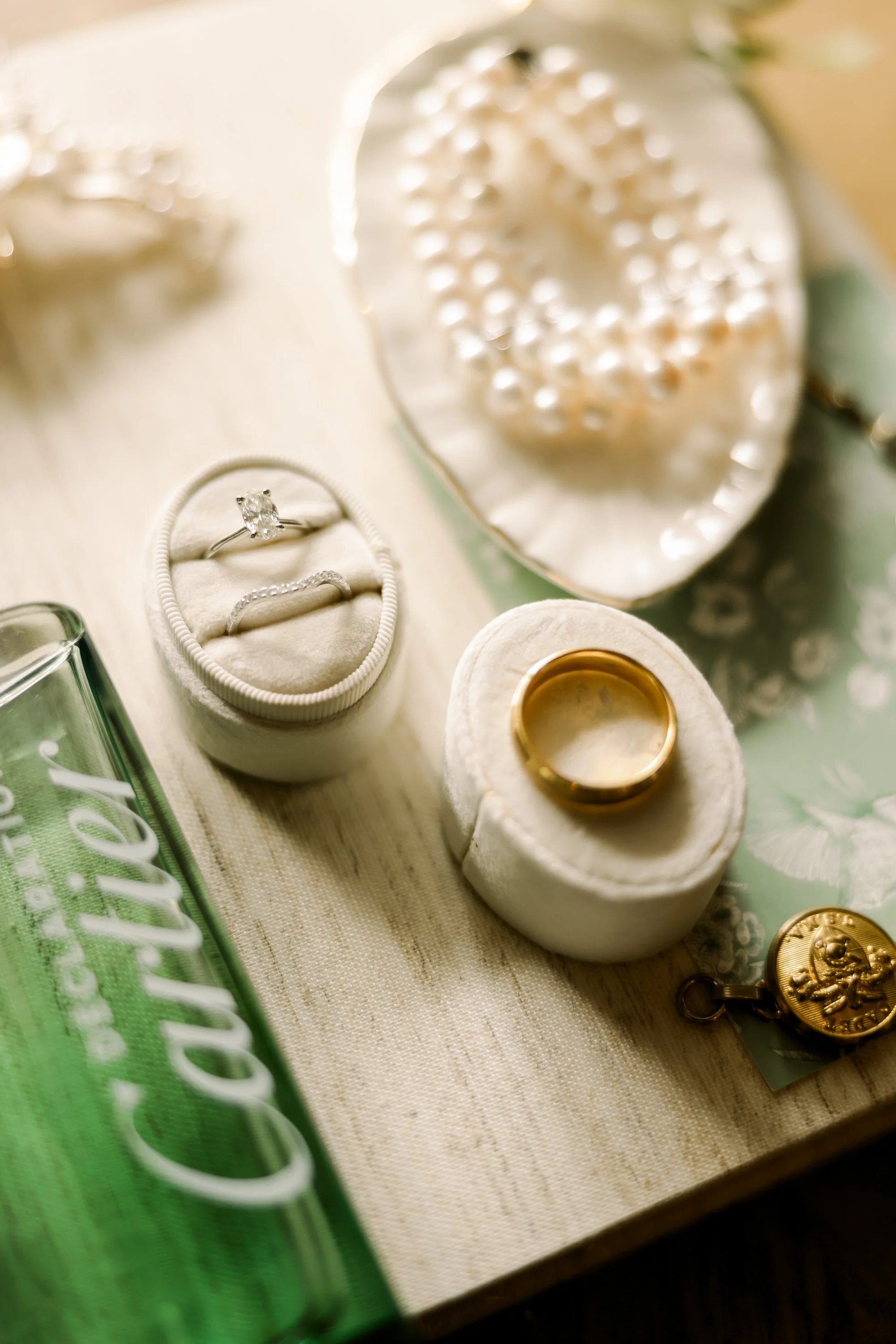 Close-up of wedding rings and jewelry on a light-colored surface, including a diamond engagement ring in a white padded box, a gold wedding band on a white cushion, a pearl necklace in a shell-shaped dish, and a gold pendant with a child's face engra