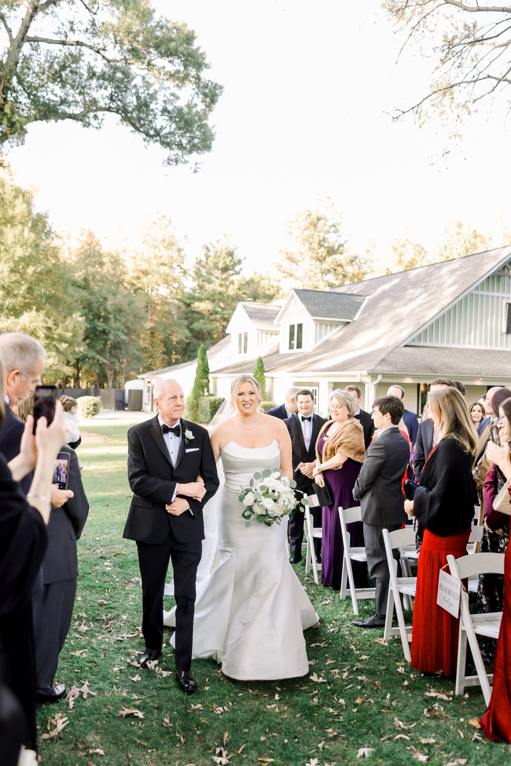 Bride walking down the aisle at an outdoor wedding ceremony with her father, surrounded by guests on a green lawn with fall leaves, in front of a house with gray siding.