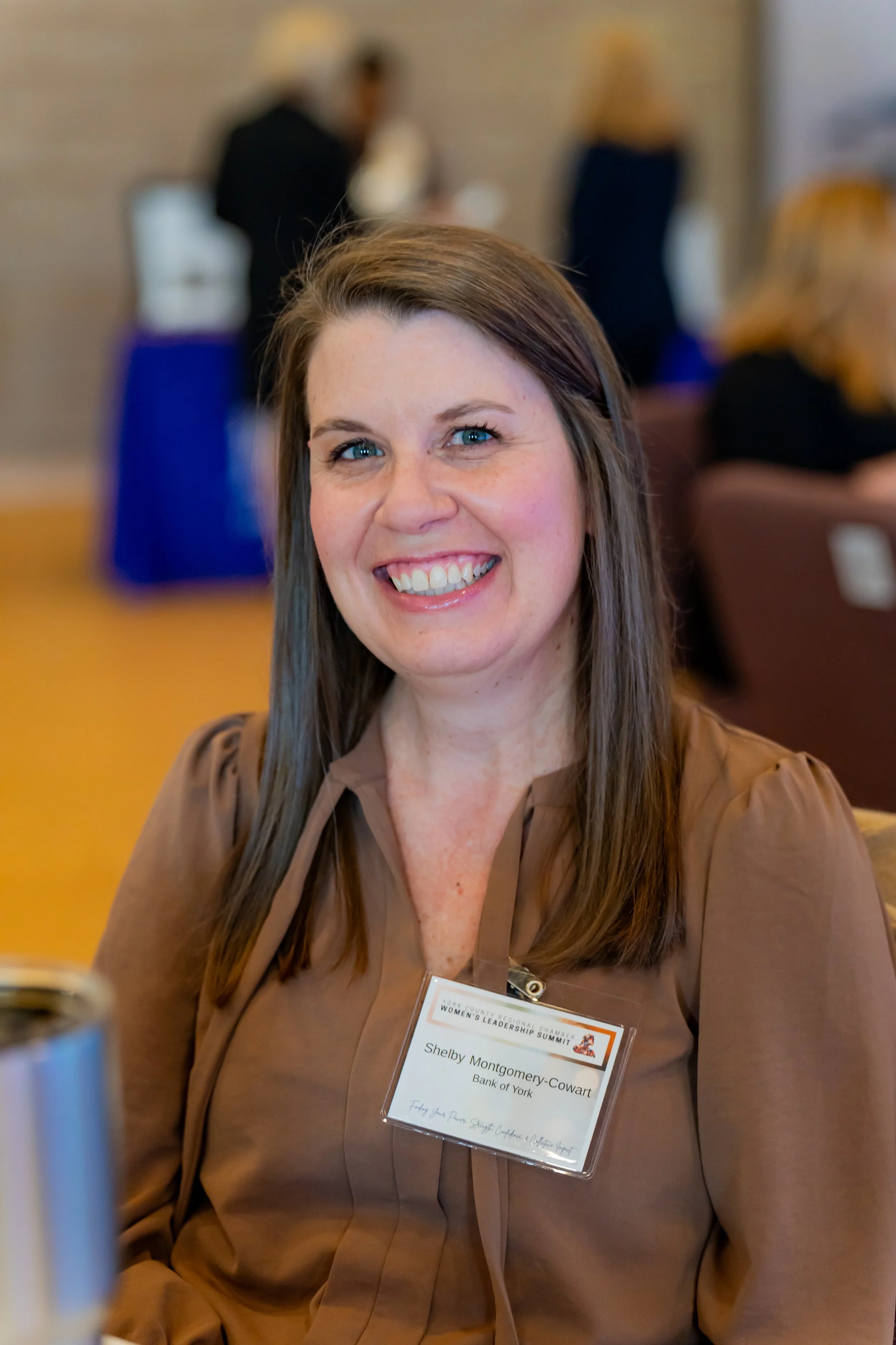 A woman with smiling face and long brown hair, wearing a brown blouse, sitting at a conference, with a name tag that reads Shelby Montgomery-Cowart, Bank of York.