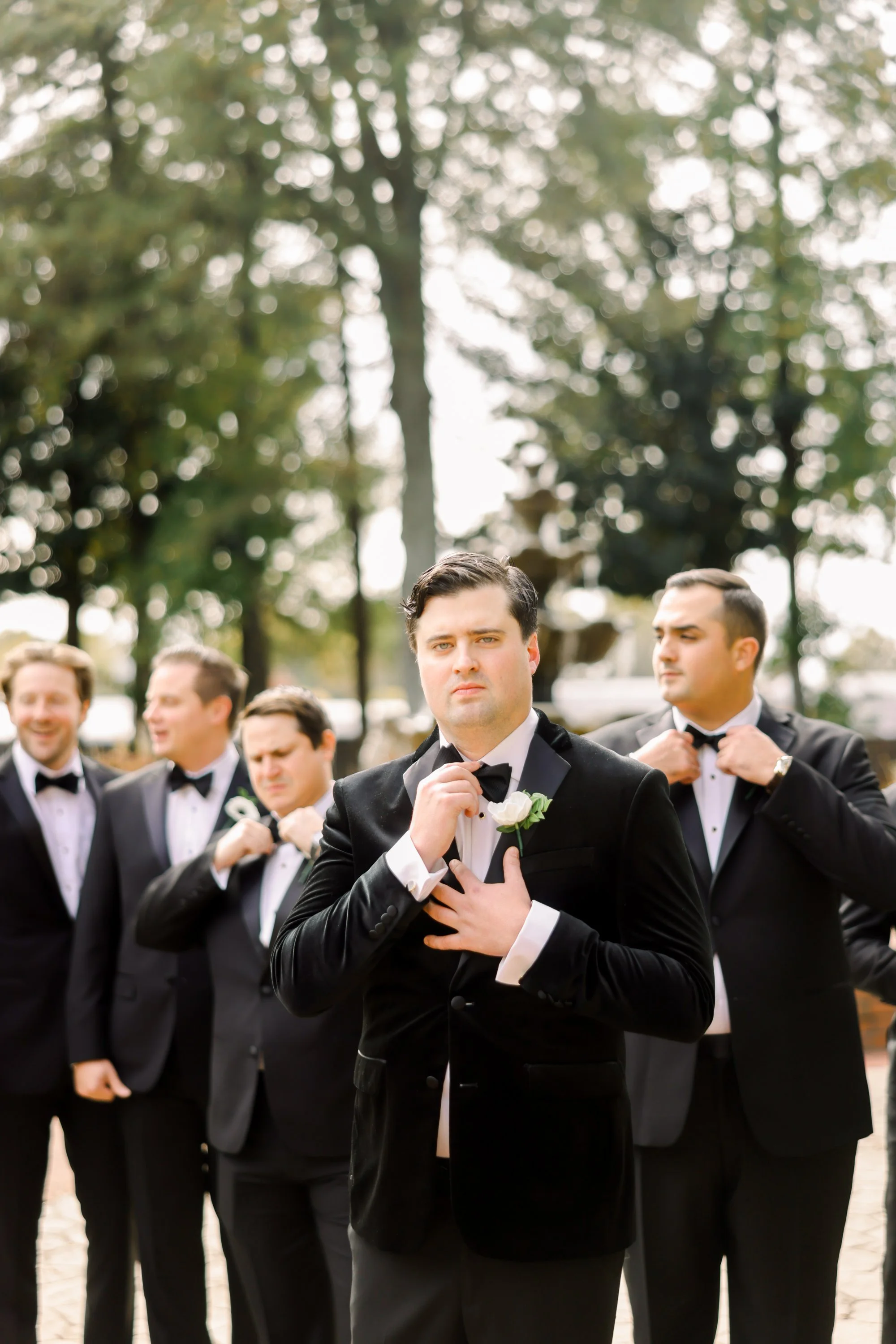 A group of five men in black tuxedos with bow ties, four of whom are adjusting their bow ties, standing outdoors with trees in the background. The central man is holding his hand to his chest, looking serious.