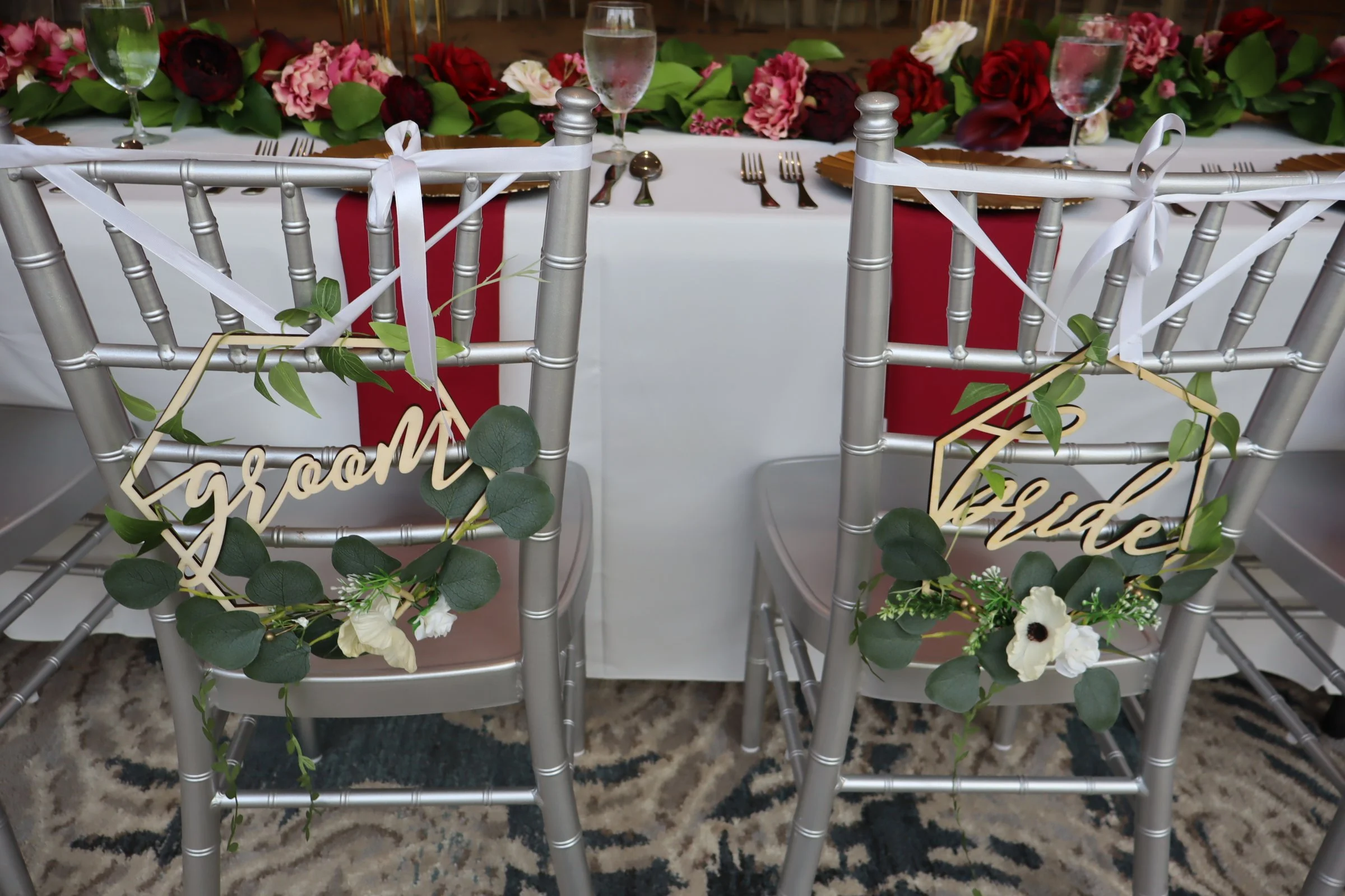 Silver chairs with signs saying 'bride' and 'groom' decorated with green leaves and white flowers, facing a decorated banquet table with floral arrangement, water glasses, and place settings.