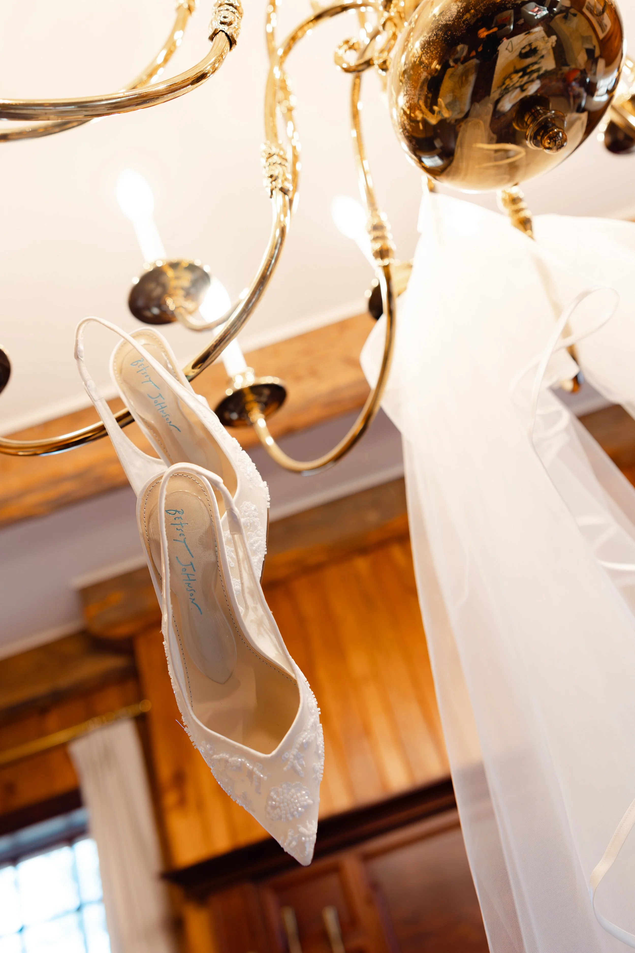 A pair of white wedding shoes with lace and floral embroidery hanging from a chandelier above a wedding dress with a flowing train.
