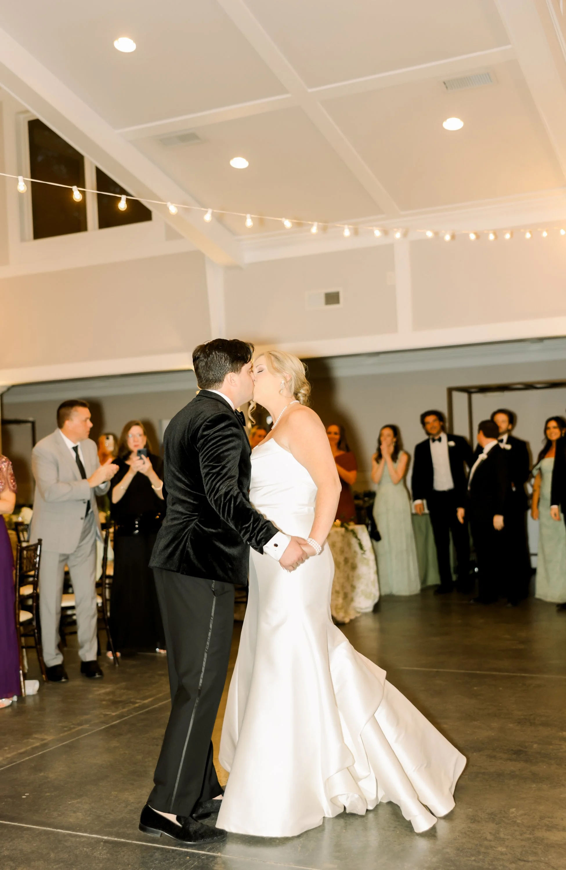 A newlywed couple sharing their first dance, surrounded by onlookers at a wedding reception, with string lights overhead in a decorated indoor venue.
