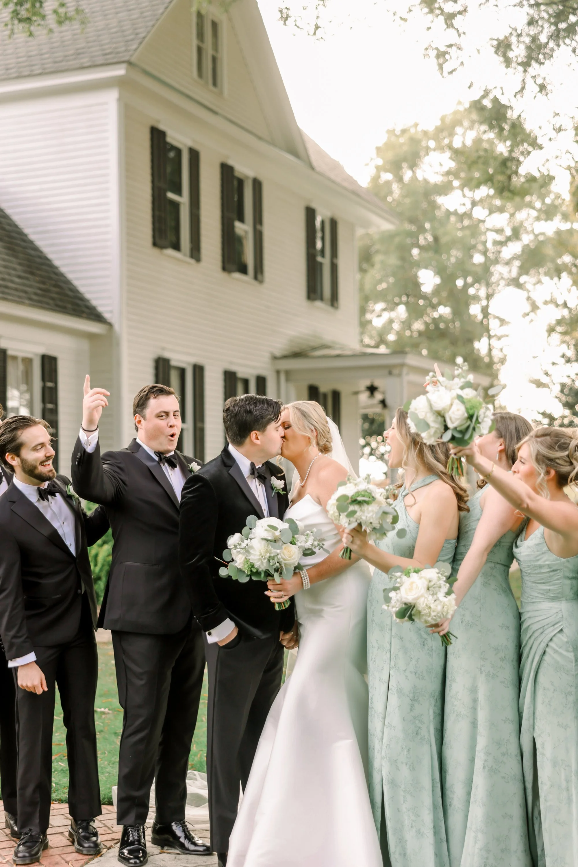 A wedding party outside a white house with black shutters, celebrating by kissing and holding flowers.