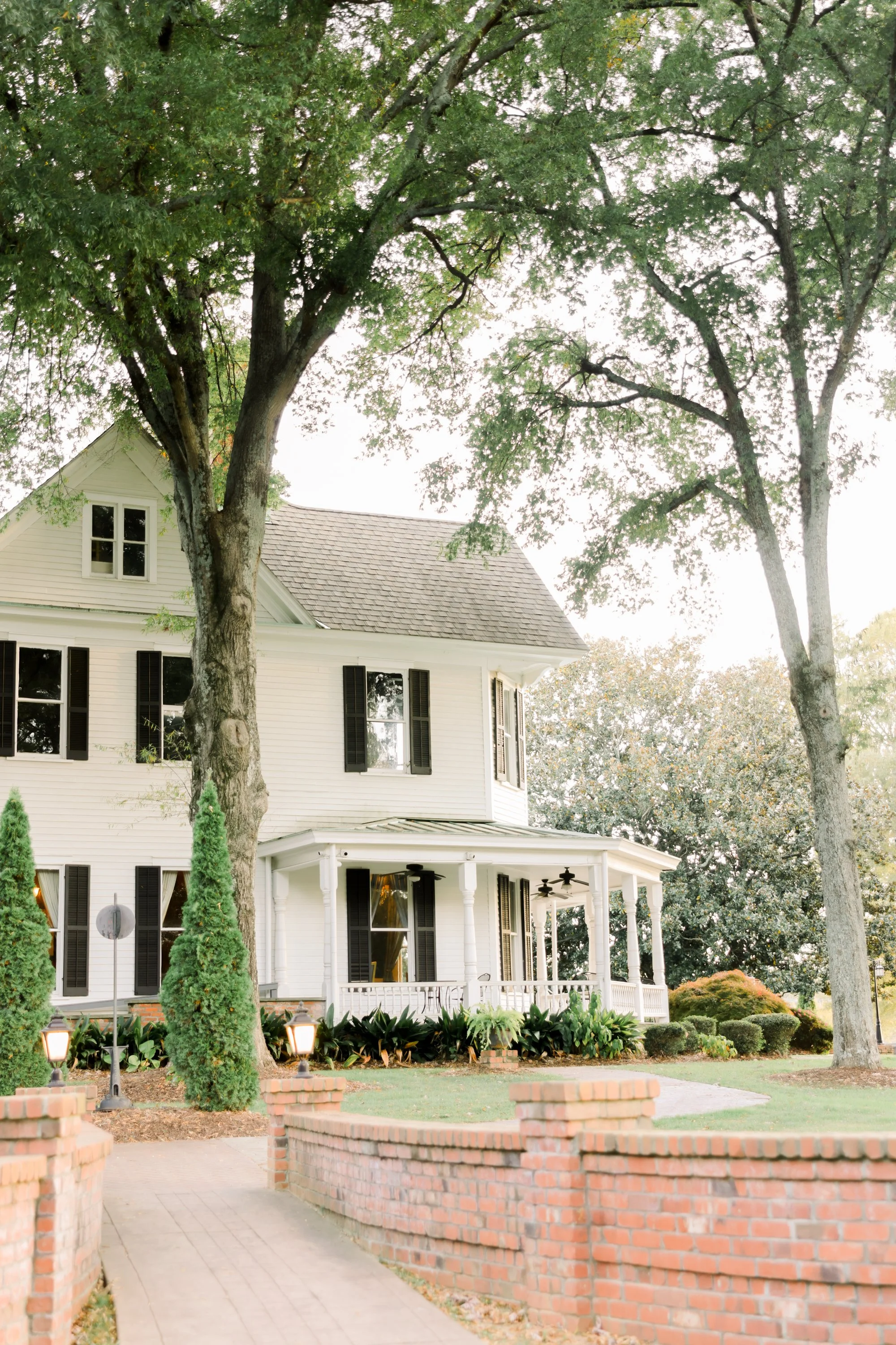 A large white house with black shutters, a covered front porch, and a garden with shrubs and trees. Two tall trees are in the foreground, and there is a brick pathway with a low brick wall leading to the house.