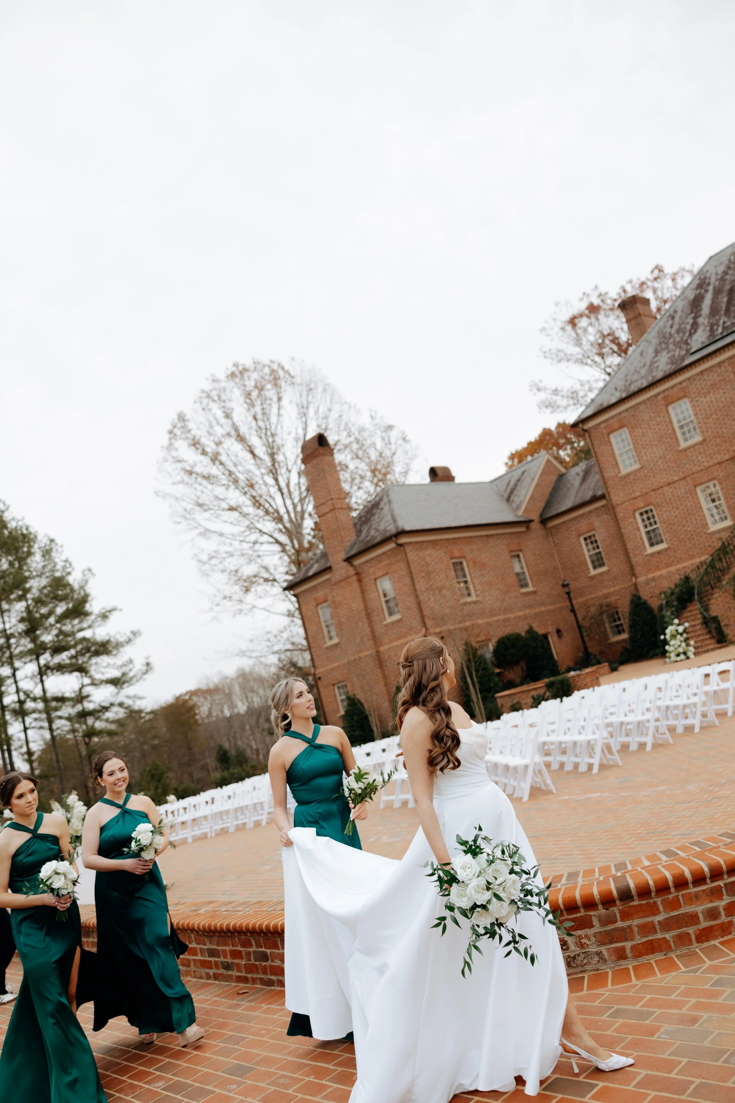 Bride in white wedding dress holding a bouquet, leading bridesmaids in teal dresses holding smaller bouquets, in an outdoor wedding setting with a brick building and white chairs in the background.