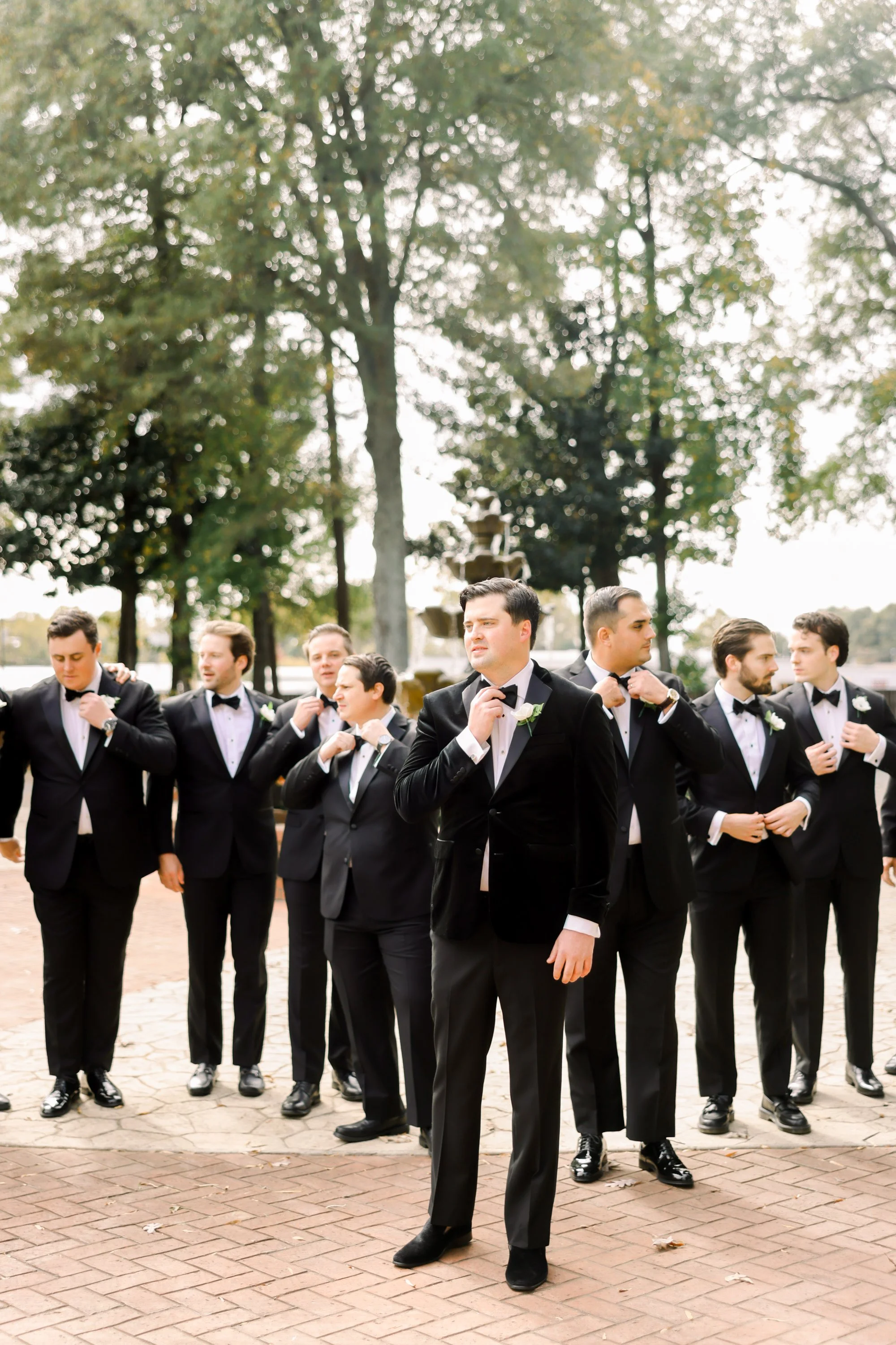 Group of men in black tuxedos adjusting their bow ties outdoors with trees in the background.