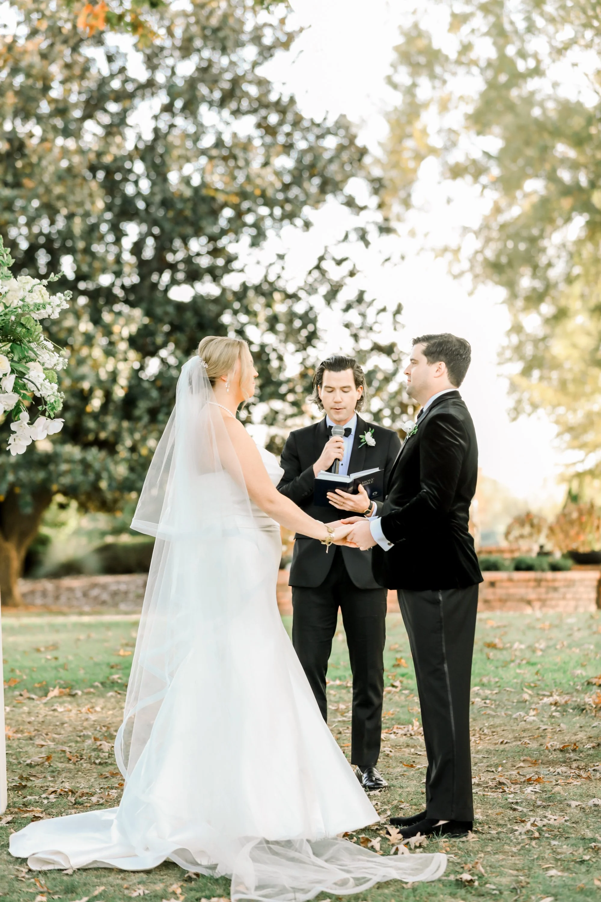 A wedding ceremony outdoors with a bride and groom holding hands, facing each other, while a officiant conducts the ceremony in front of large trees and greenery.