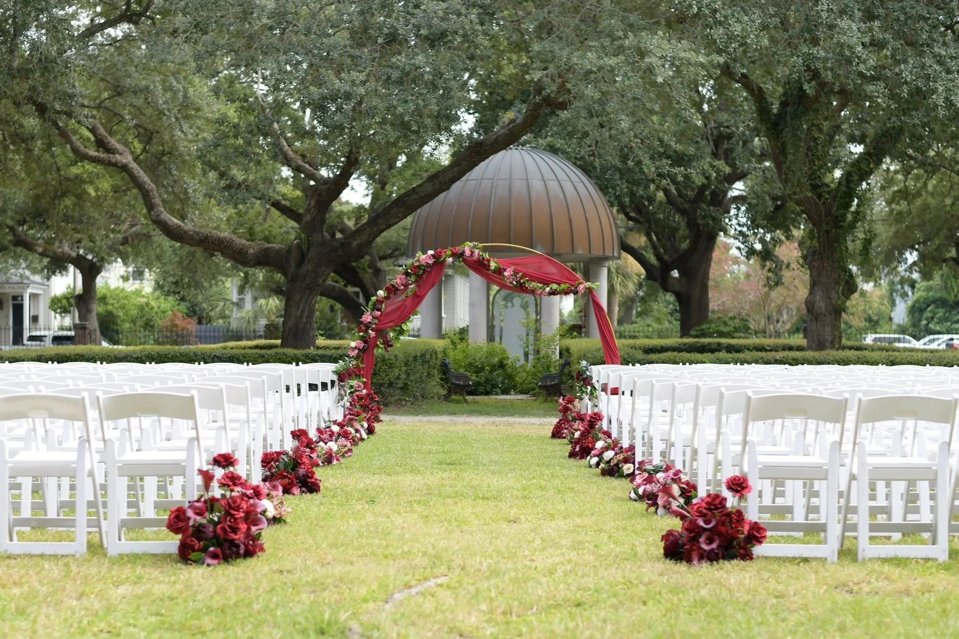 Outdoor wedding ceremony setup with white chairs arranged in two rows, decorated with red and pink floral arrangements along the aisle, centered by a floral arch with red fabric drapes, in a green park with large trees and a small pavilion in the bac