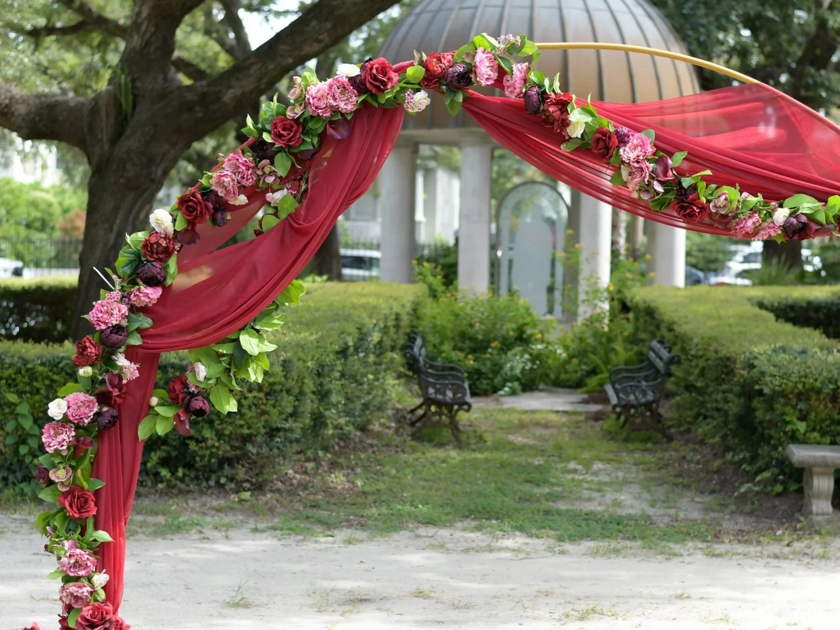 Decorative garden arch with pink and red flowers and sheer pink fabric, set in a park with benches, trees, and a structure with columns