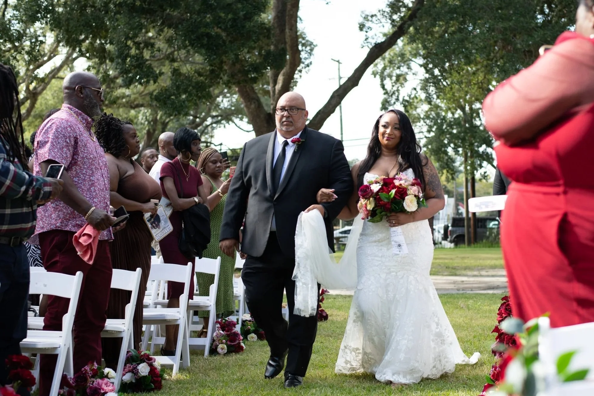 A bride walking down the aisle with her escort during an outdoor wedding ceremony. She is holding a bouquet of red and white flowers, and the guests are watching and taking pictures.