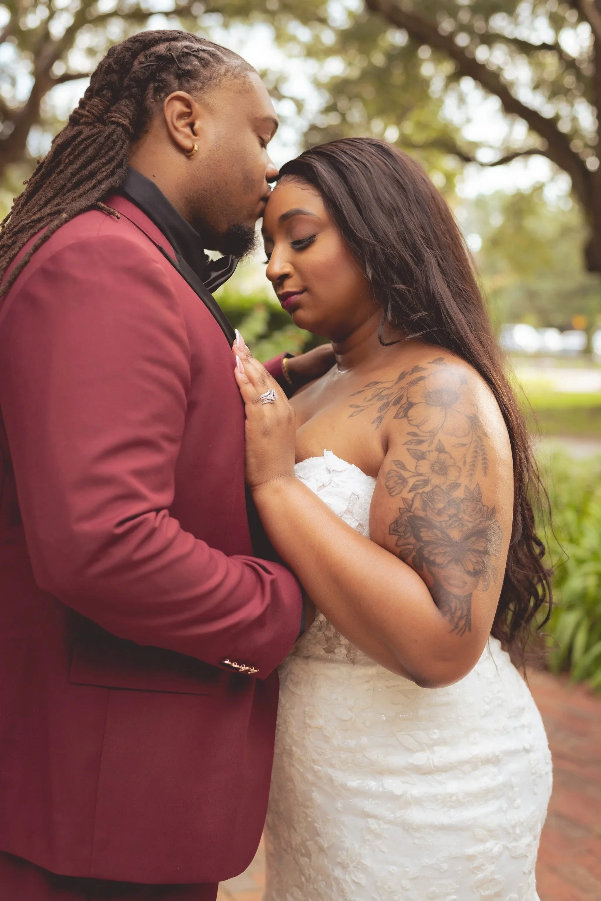 A couple dressed in wedding attire sharing an intimate moment outdoors, with the man in a maroon suit and the woman in a white lace wedding dress, both with their eyes closed and touching foreheads.