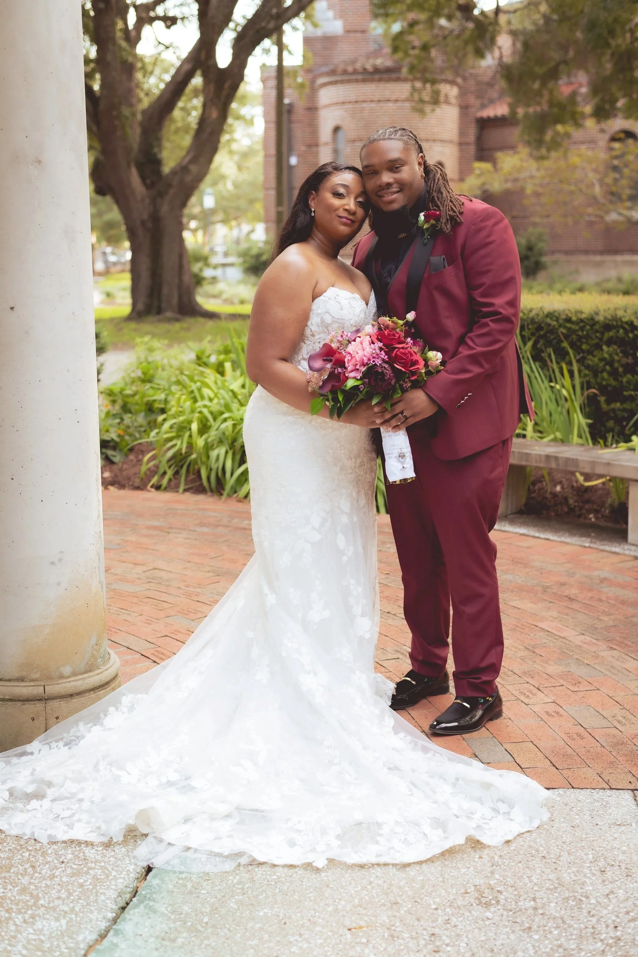 A newlywed couple poses outdoors, with the bride in a white lace wedding gown holding a colorful bouquet, and the groom in a burgundy suit with a boutonnière, standing close together and smiling.