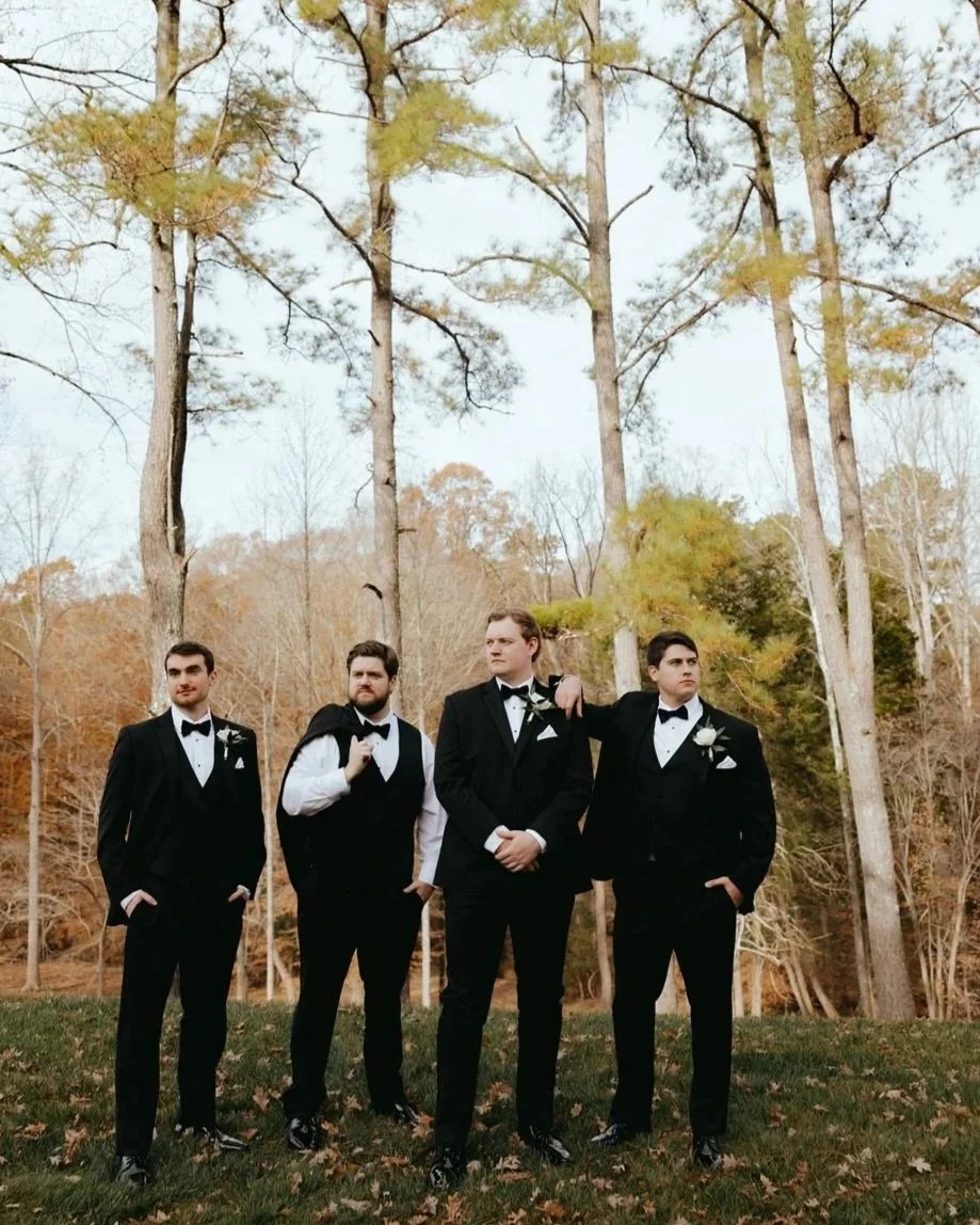Four men in tuxedos standing outdoors on grass with trees in fall colors behind them.