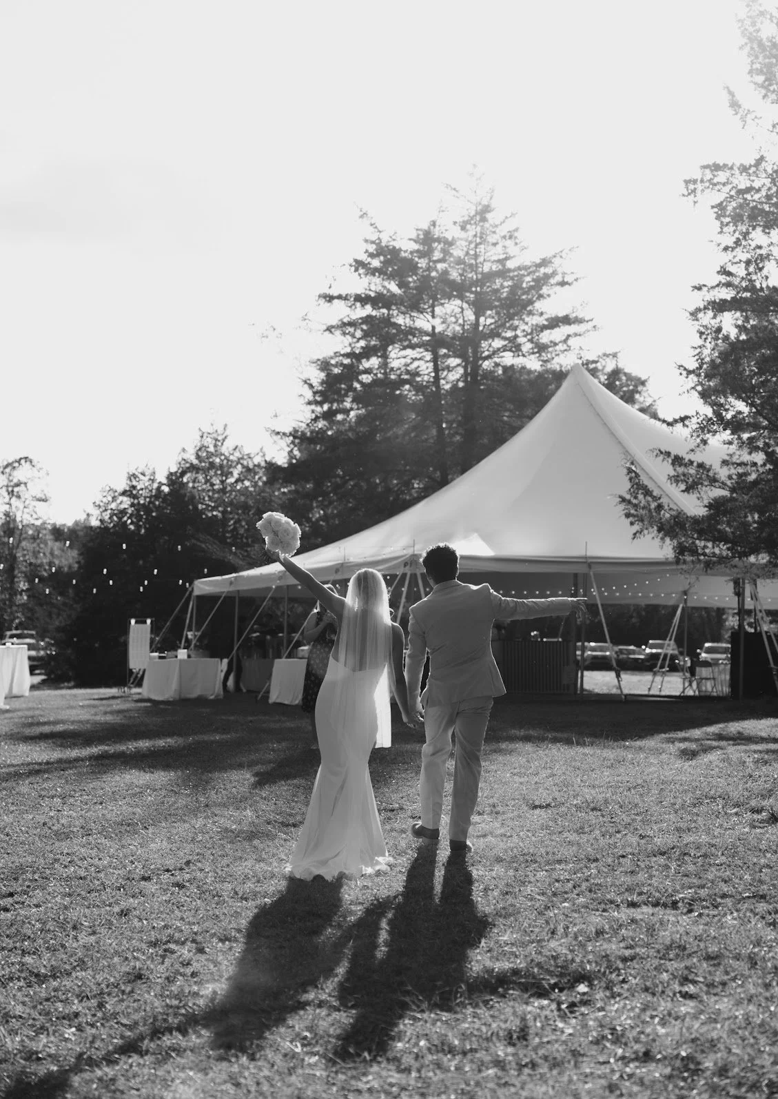 A black and white photo of a bride and groom holding hands and walking away from a large tent at an outdoor wedding reception, with the bride holding a bouquet and the groom in a suit.