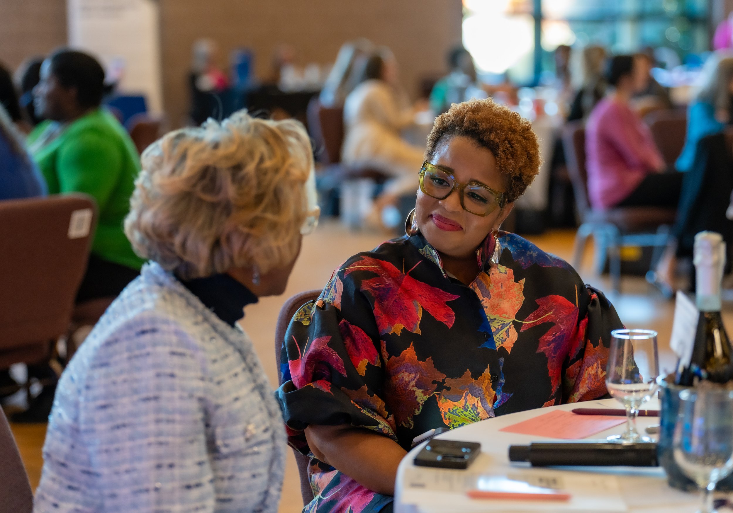Two women engaged in conversation at a conference, sitting at a round table with a glass of water and a smartphone on it, in a room filled with other attendees.