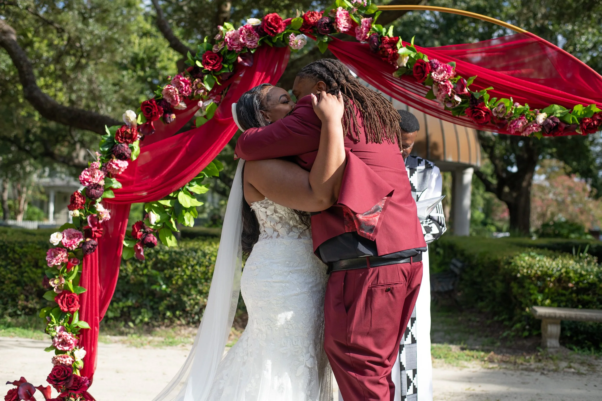 A couple hugging at their outdoor wedding ceremony, under a red floral arch with pink and red flowers. The bride is in a white wedding dress, and the groom is wearing a maroon suit. A man in a black and white patterned outfit stands nearby, and trees and a building are in the background.